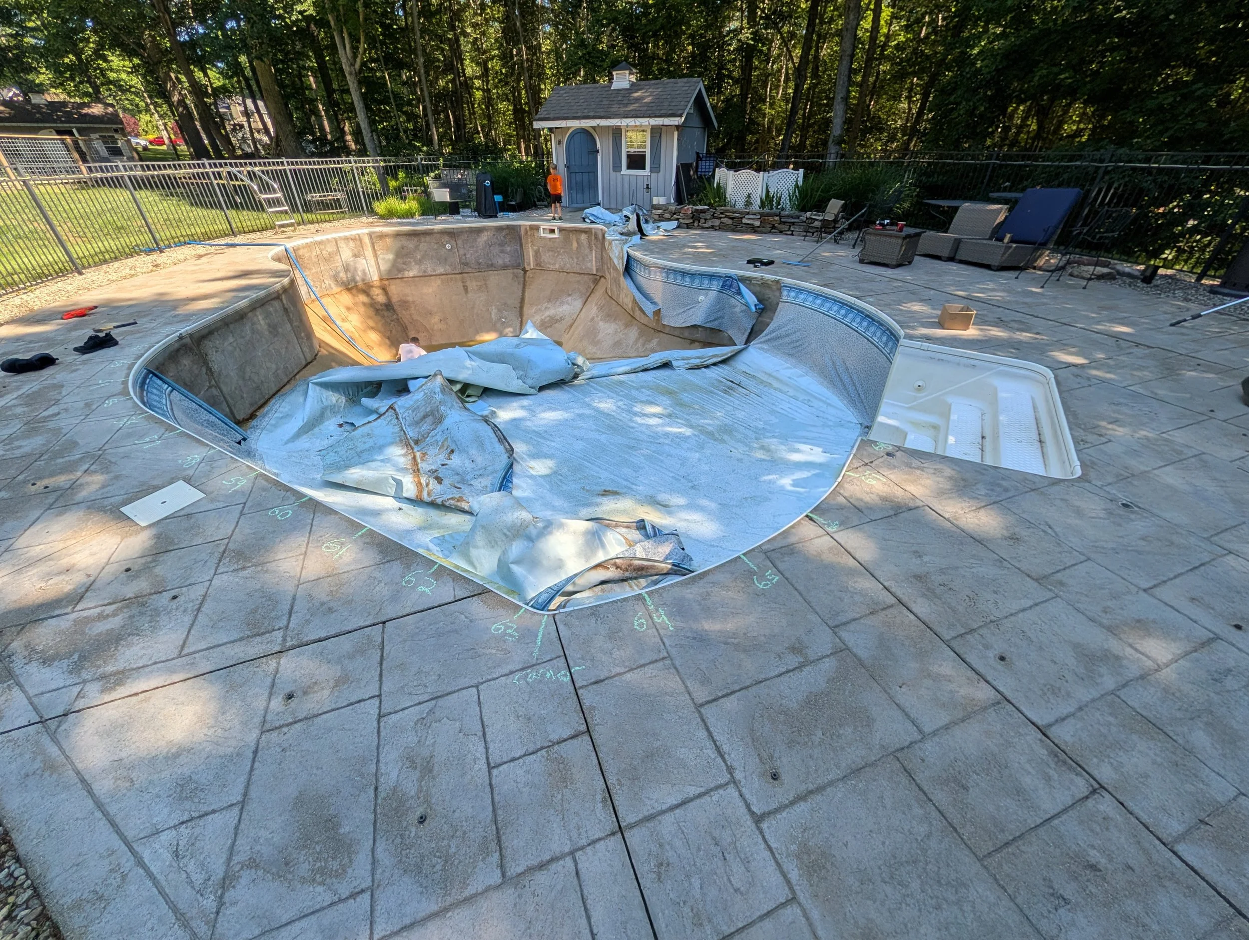 An empty in-ground swimming pool being renovated with a cover partially peeled back, surrounded by outdoor patio furniture, a small shed, and a backyard with trees.