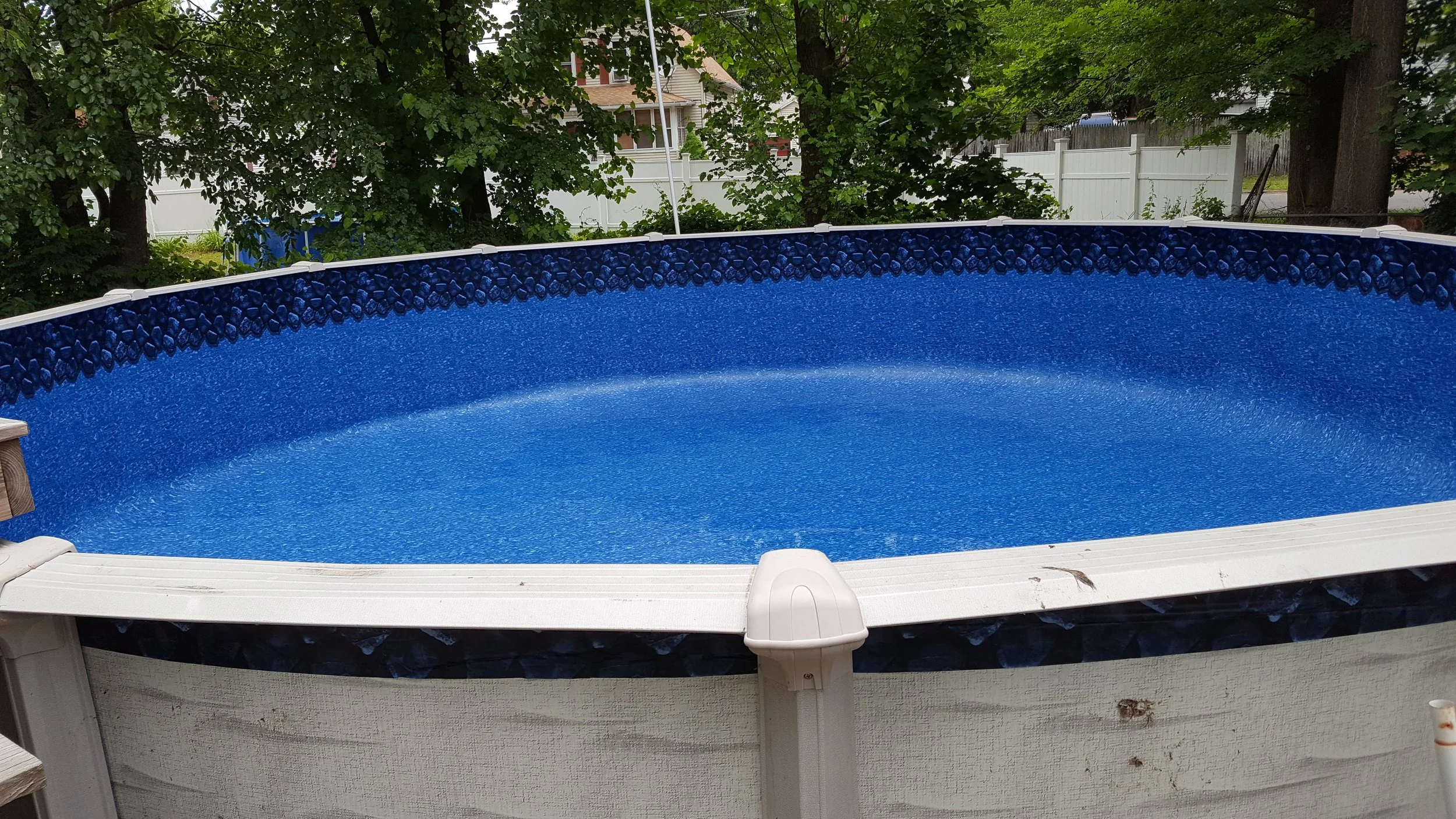 Above-ground oval swimming pool with blue interior and a decorative black rock-like border, situated outdoors in a backyard with trees, a white fence, and neighboring houses in the background.