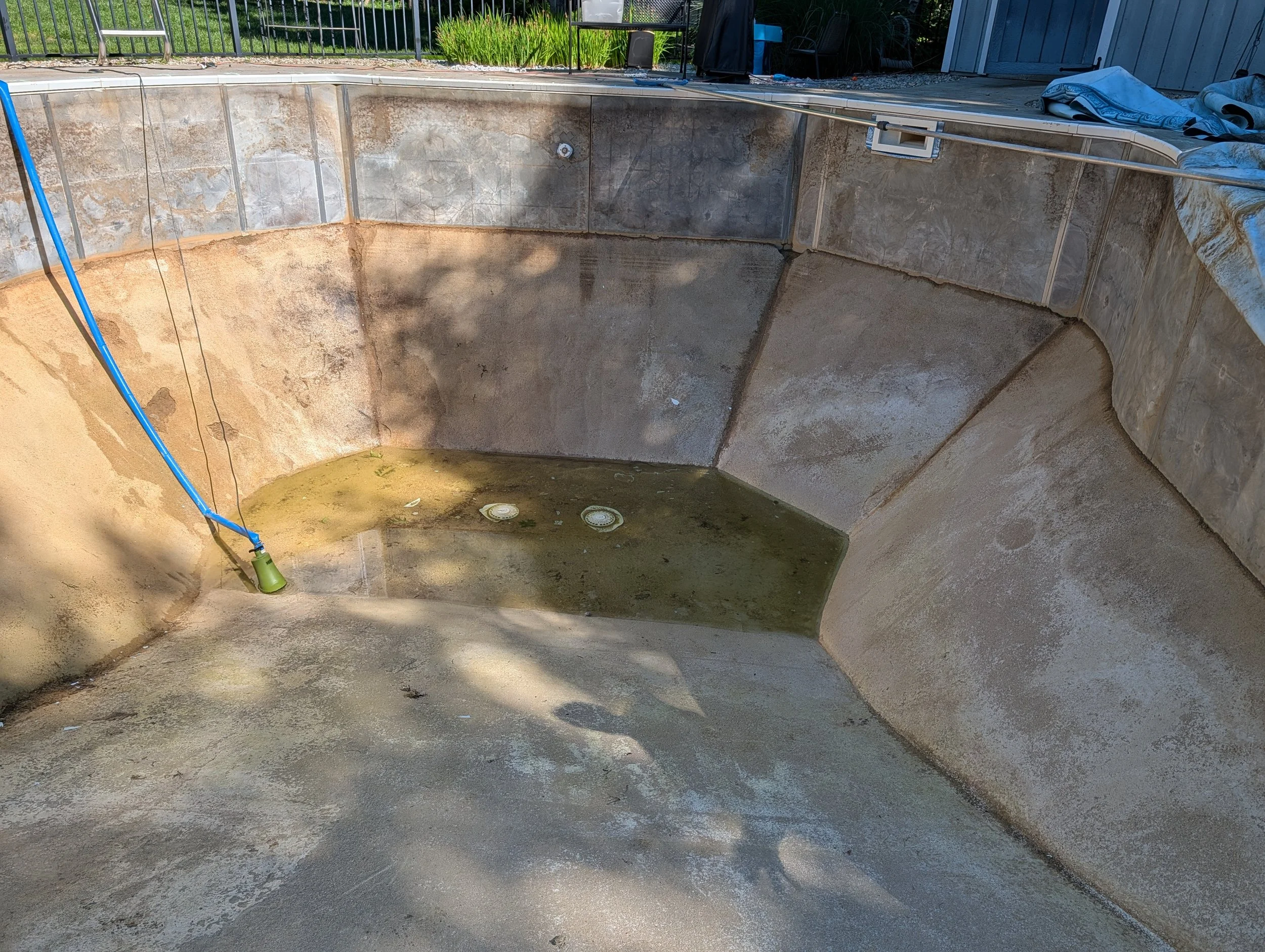 An empty swimming pool with a sandy bottom and tiled walls, surrounded by some pool equipment and a fence, with a house and green plants in the background.