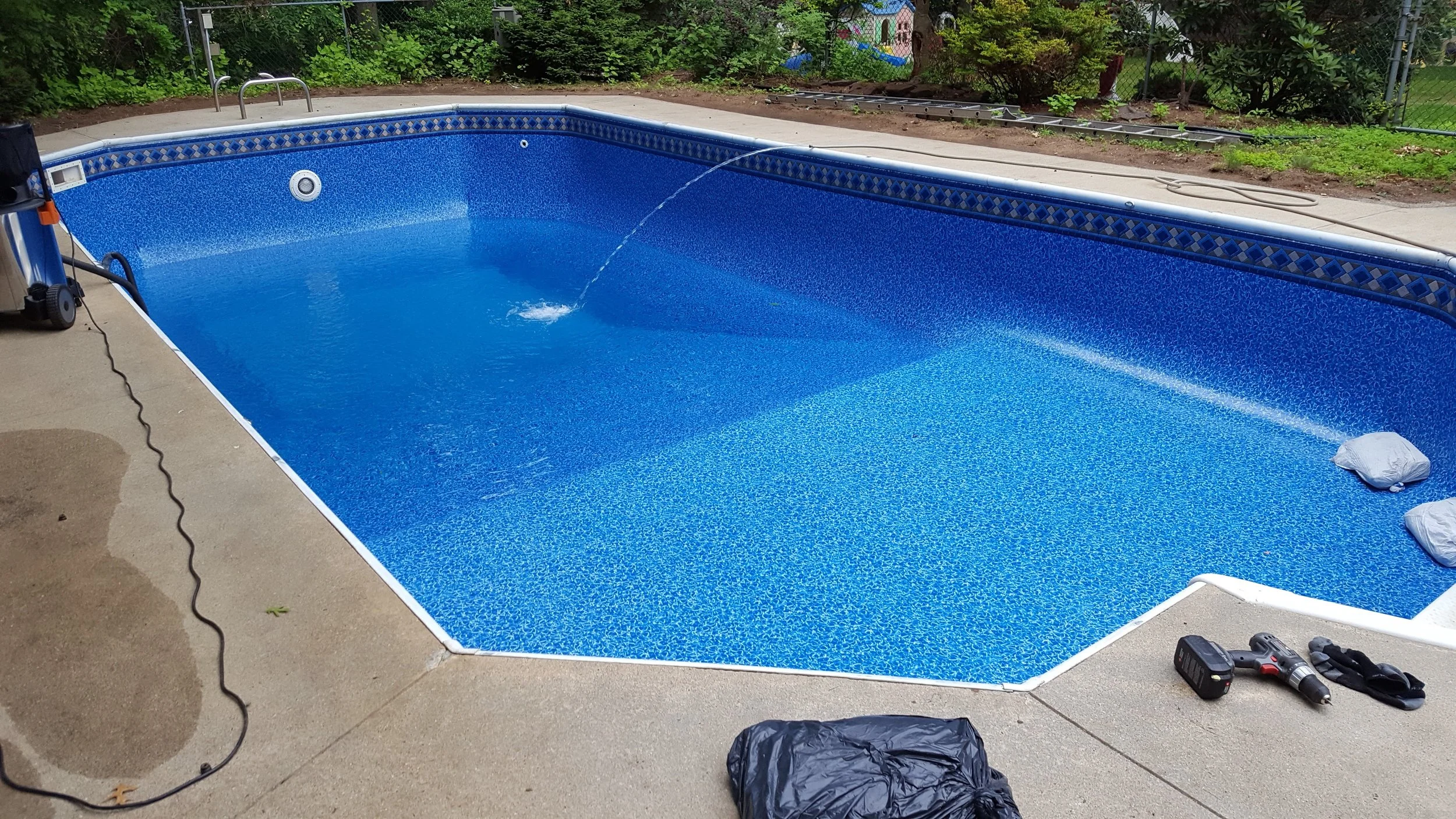 Empty in-ground swimming pool with blue tiles, pool cover folded at one end, surrounded by concrete patio, with tools and supplies nearby, in a backyard with trees and a chain-link fence.