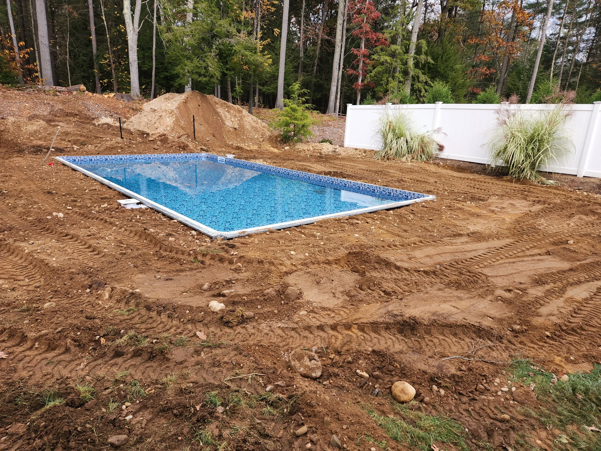 Under construction or landscaping area with dirt, rocks, and a small pile of loose soil next to a white fence with tall grass plants. Forested area with trees in fall colors in the background.