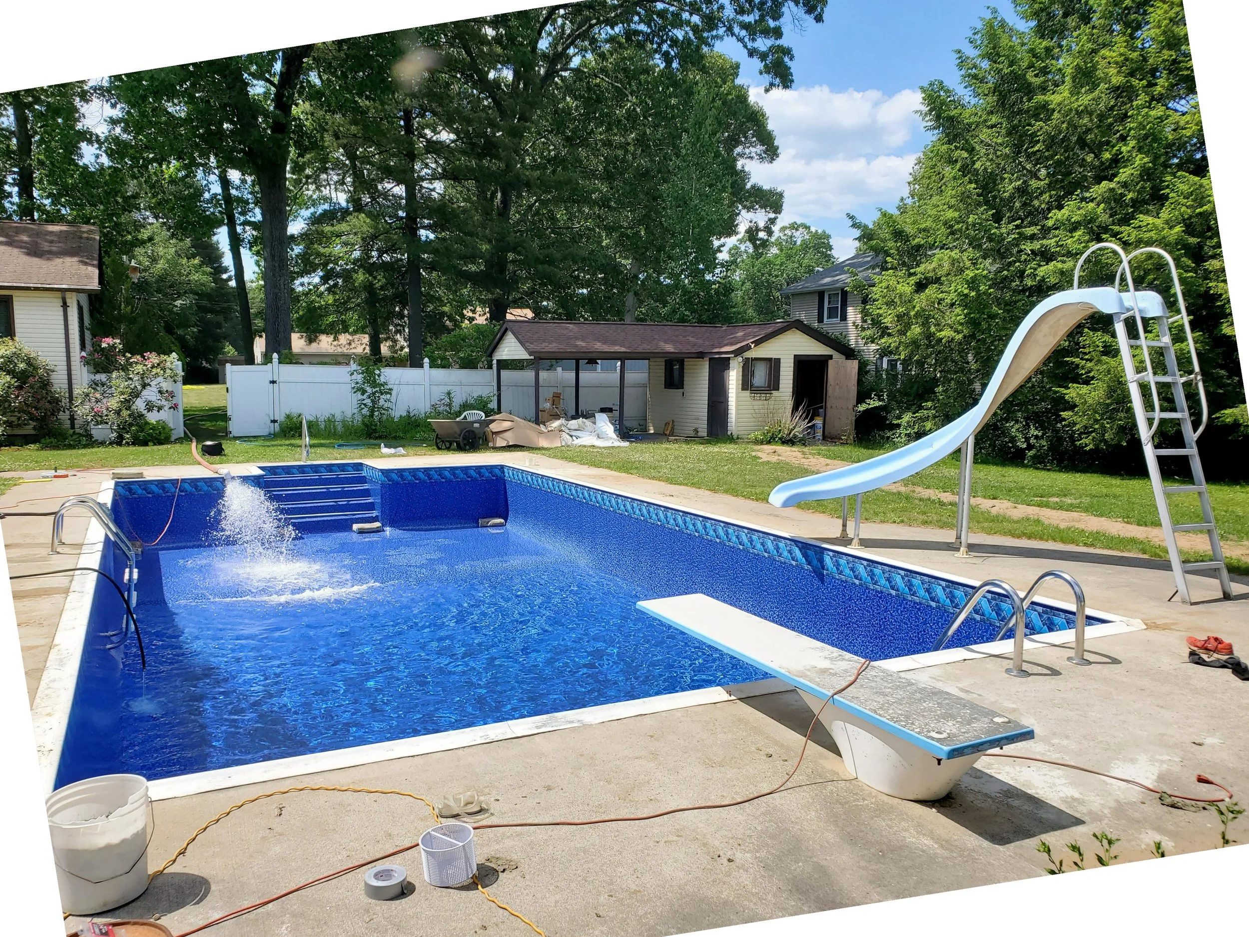 A backyard with an in-ground swimming pool and a waterslide. There is a small shed or pool house with a brown roof, and a white fence surrounding the yard. Tall trees with green leaves are in the background, and the sky is partly cloudy.