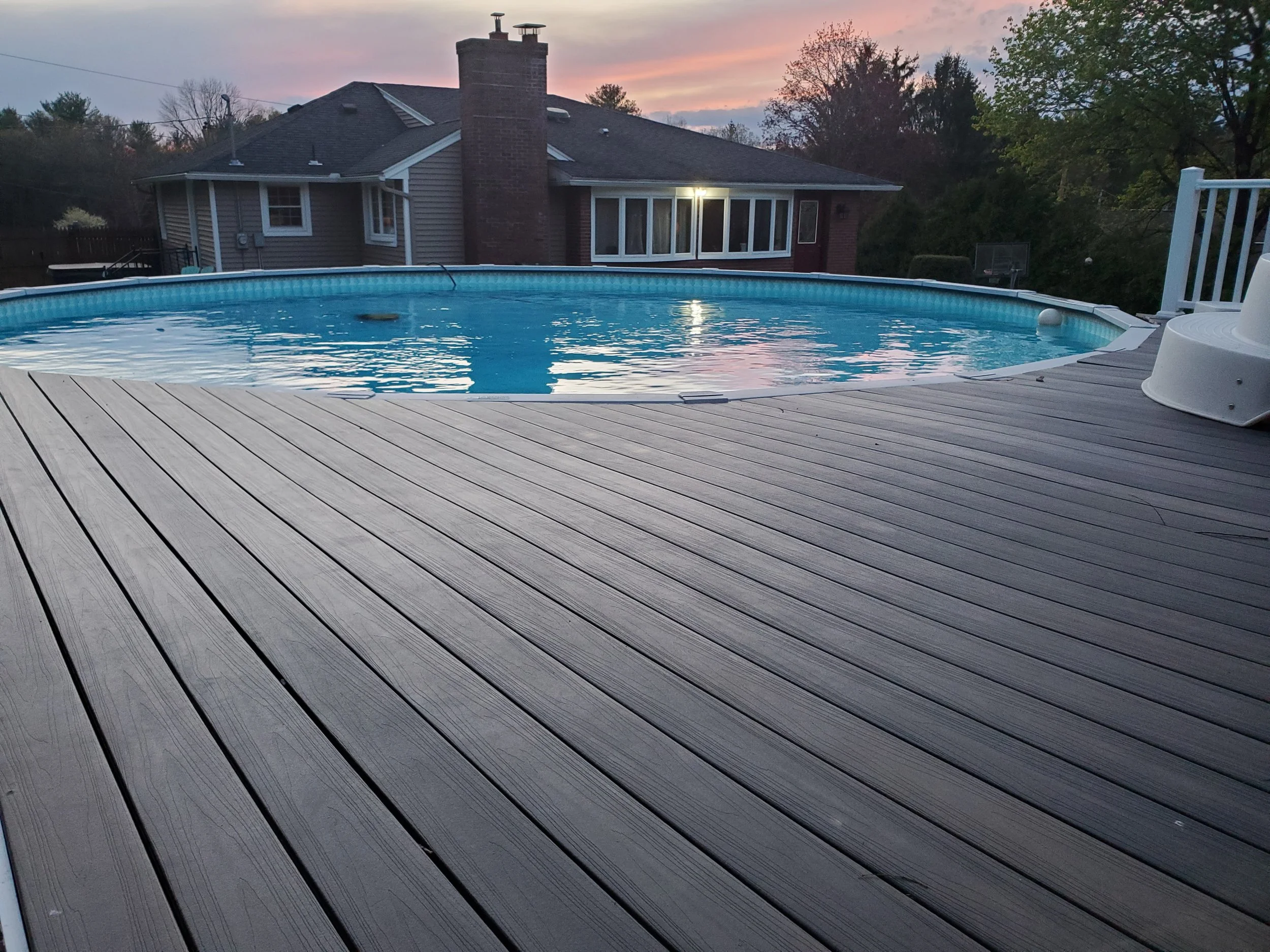 Backyard with a swimming pool, wooden deck, house in the background, trees, and a colorful sunset sky.