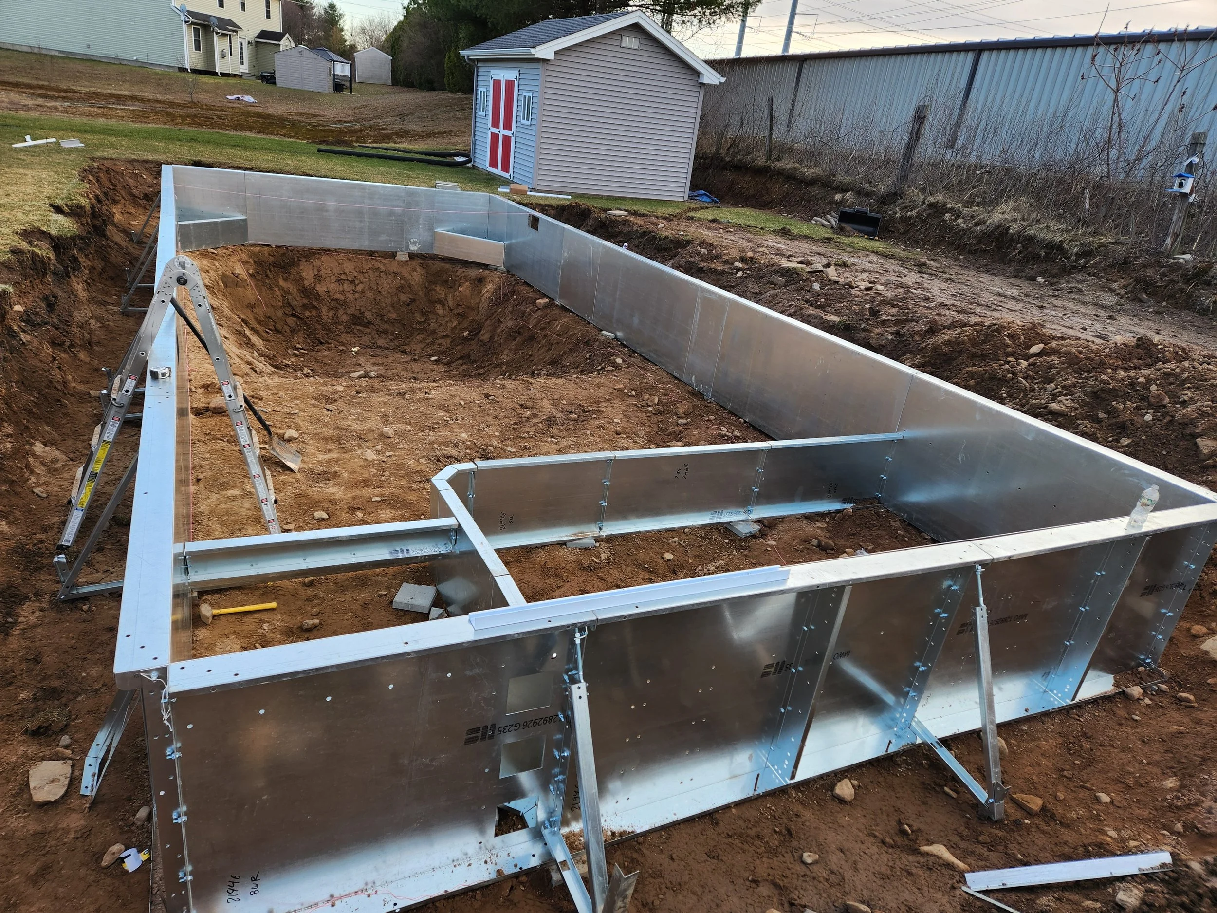 A construction site where a swimming pool is being installed, showing metal pool walls assembled in an excavation area in a backyard.