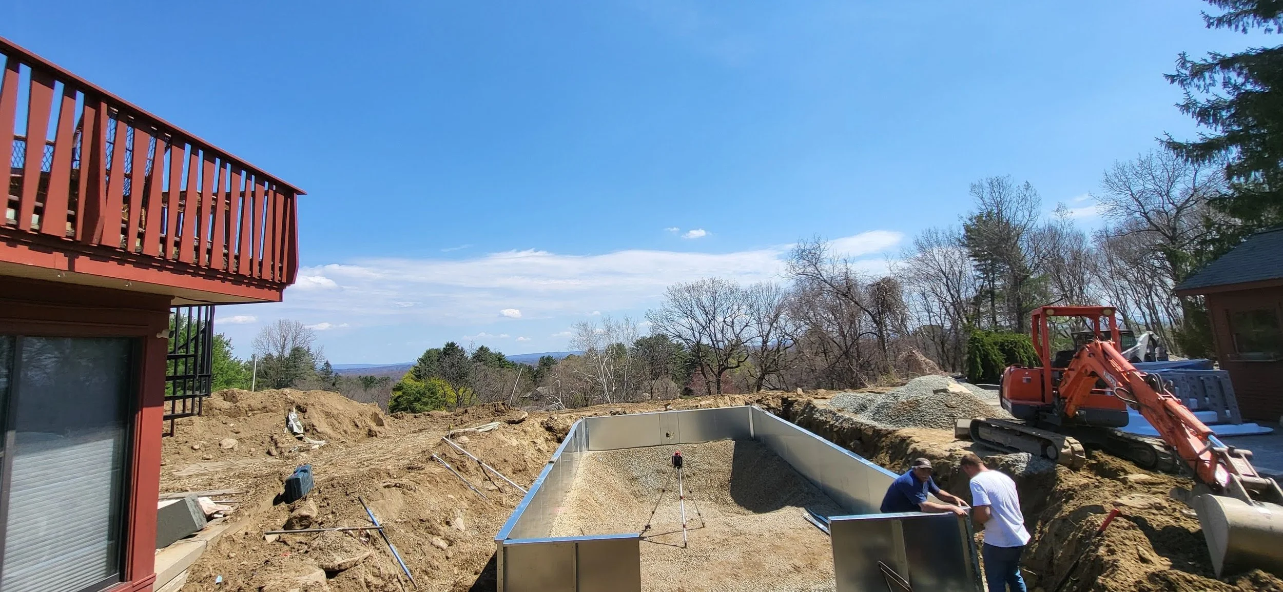 Construction site with dug-out area, a small excavator, and workers, with a background of trees and a partly cloudy sky.