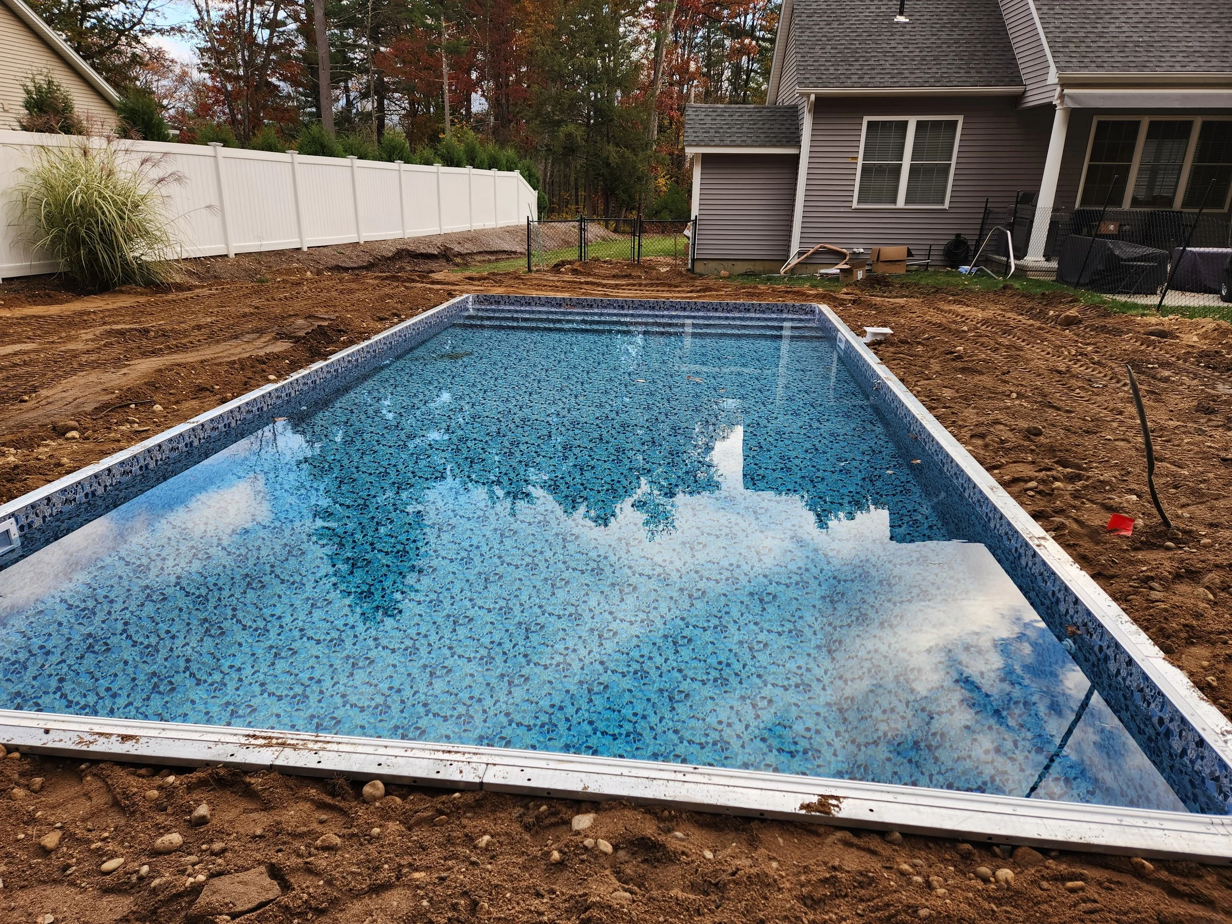 A backyard in the process of renovation with a newly installed in-ground swimming pool, surrounded by dirt and construction materials. There is a white privacy fence on the left, a house with gray siding on the right, and trees with autumn foliage in