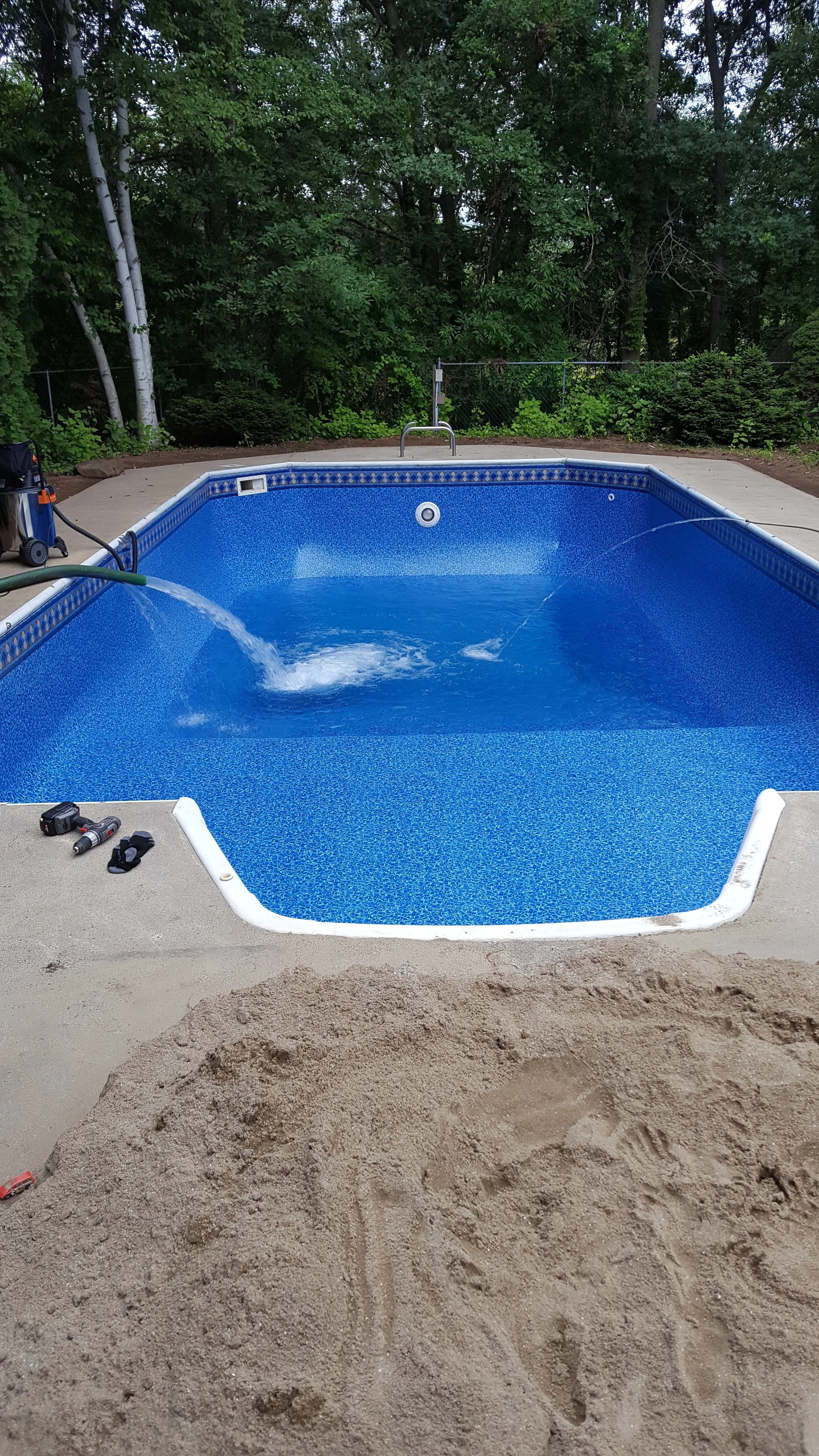 A blue in-ground swimming pool with a concrete deck, surrounded by a wooded backyard. There are tools and equipment on the pool deck, and the pool appears to be under maintenance or cleaning.