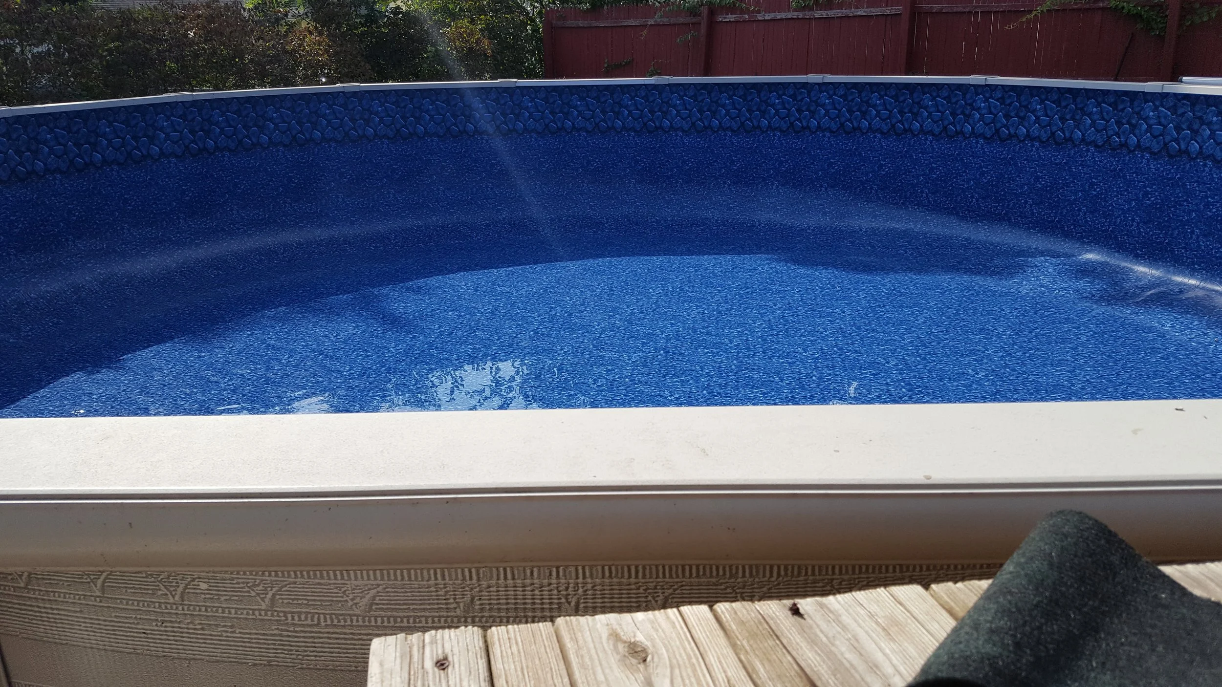 Empty blue swimming pool with a pebble border, surrounded by a red fence and trees in the background.