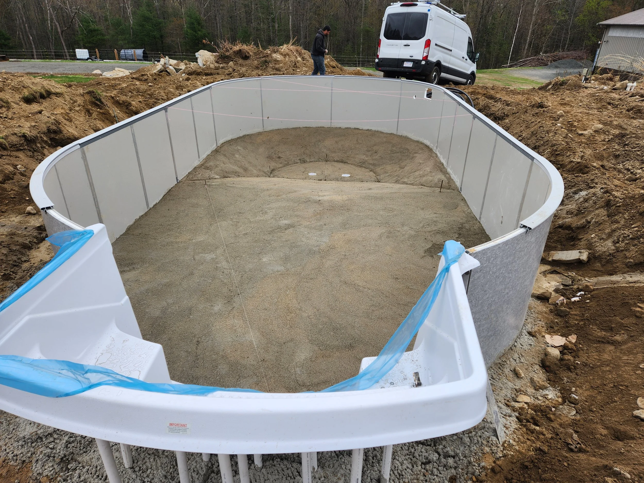 A backyard swimming pool during construction with sand inside, surrounded by dirt and construction materials, and a worker in the background near a white van.