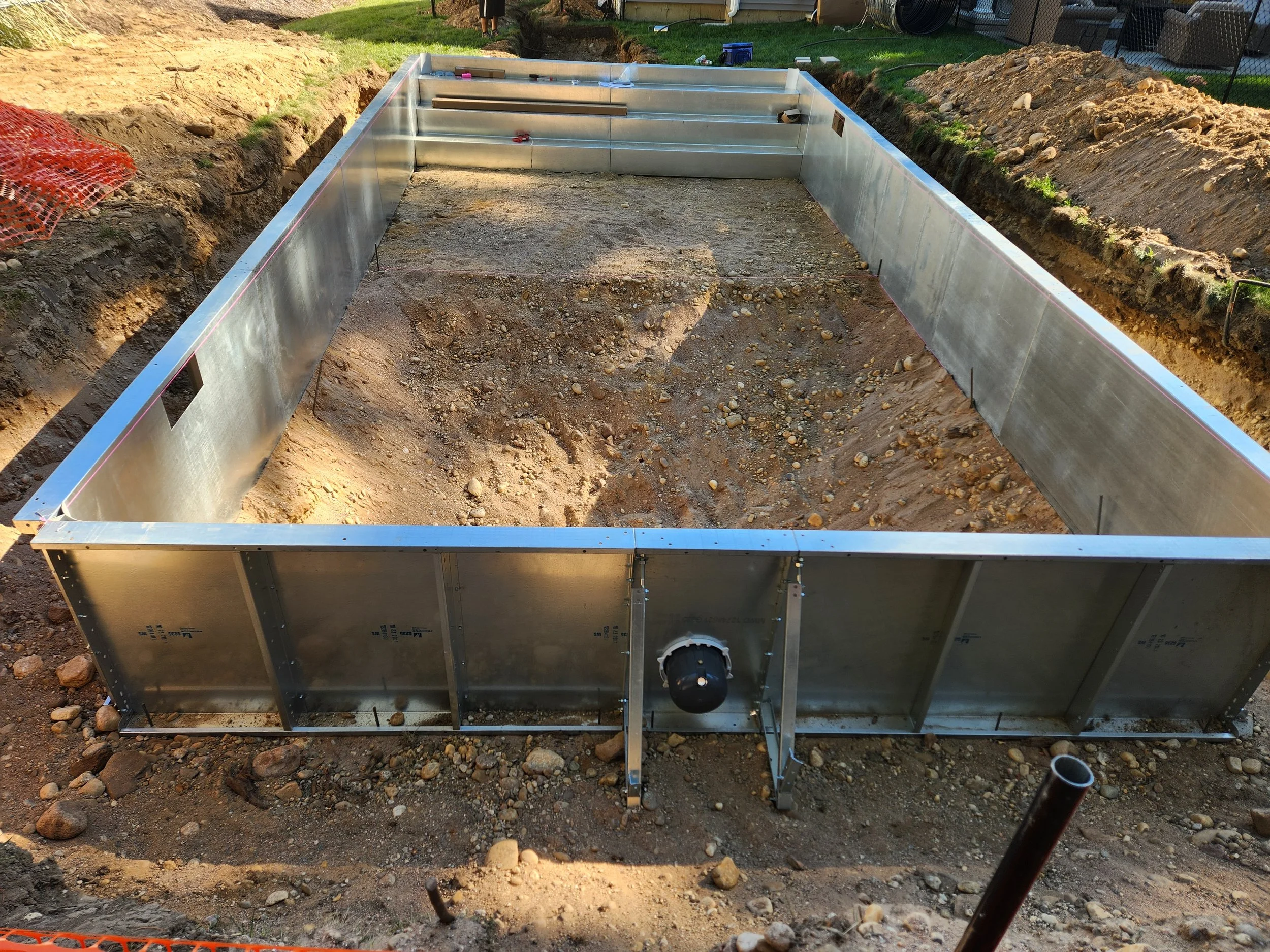 A rectangular metal pool structure is under construction in an outdoor yard, surrounded by dirt and rocks, with construction tools on top.