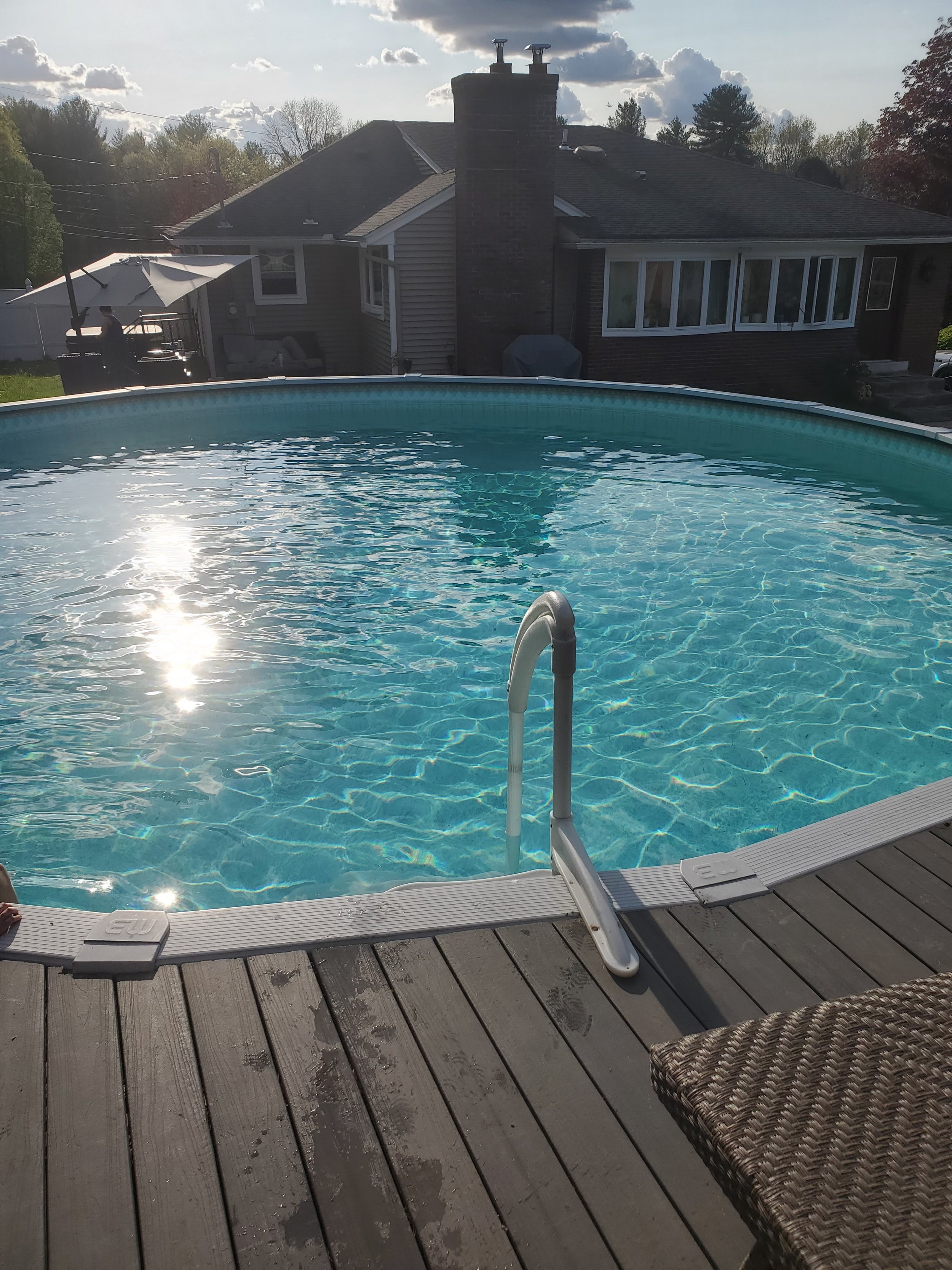 Backyard scene with a swimming pool, a wooden deck, wicker chair, an umbrella, house with large window and chimney, trees, and a partly cloudy sky.
