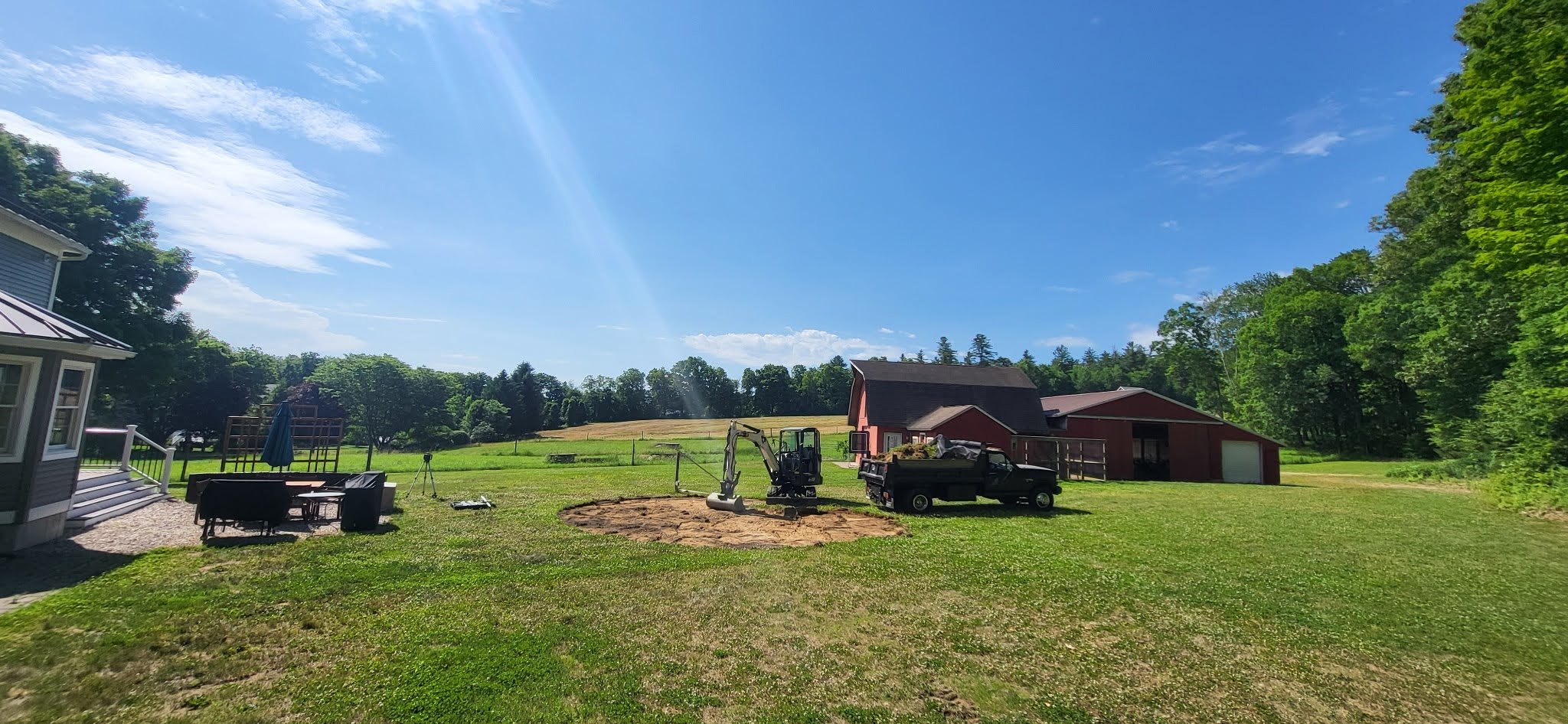 A backyard with green grass, a small excavator and truck, a red barn, and a patio with outdoor furniture under a partly cloudy blue sky.