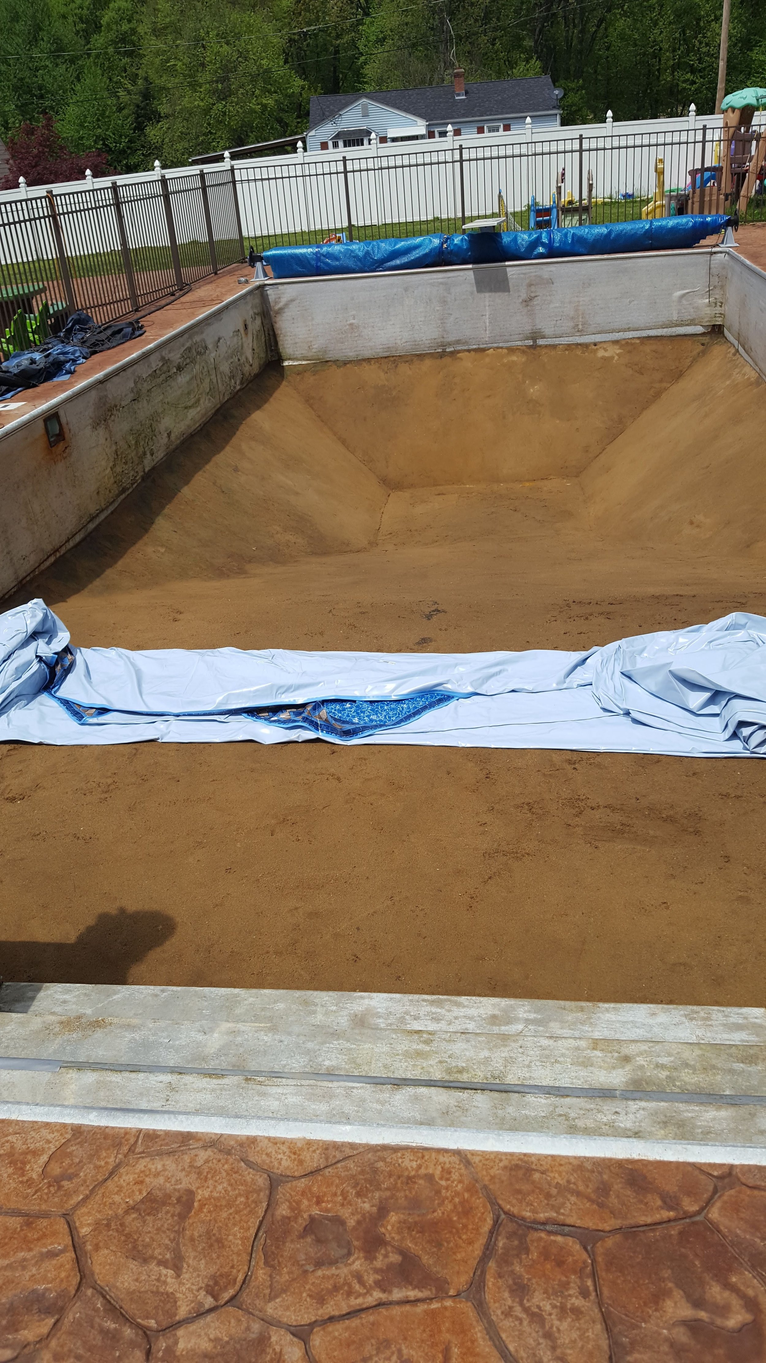 Empty swimming pool with dirt and sand inside, covering part of the pool with a white plastic sheet, located in a backyard with a white fence and green trees in the background.