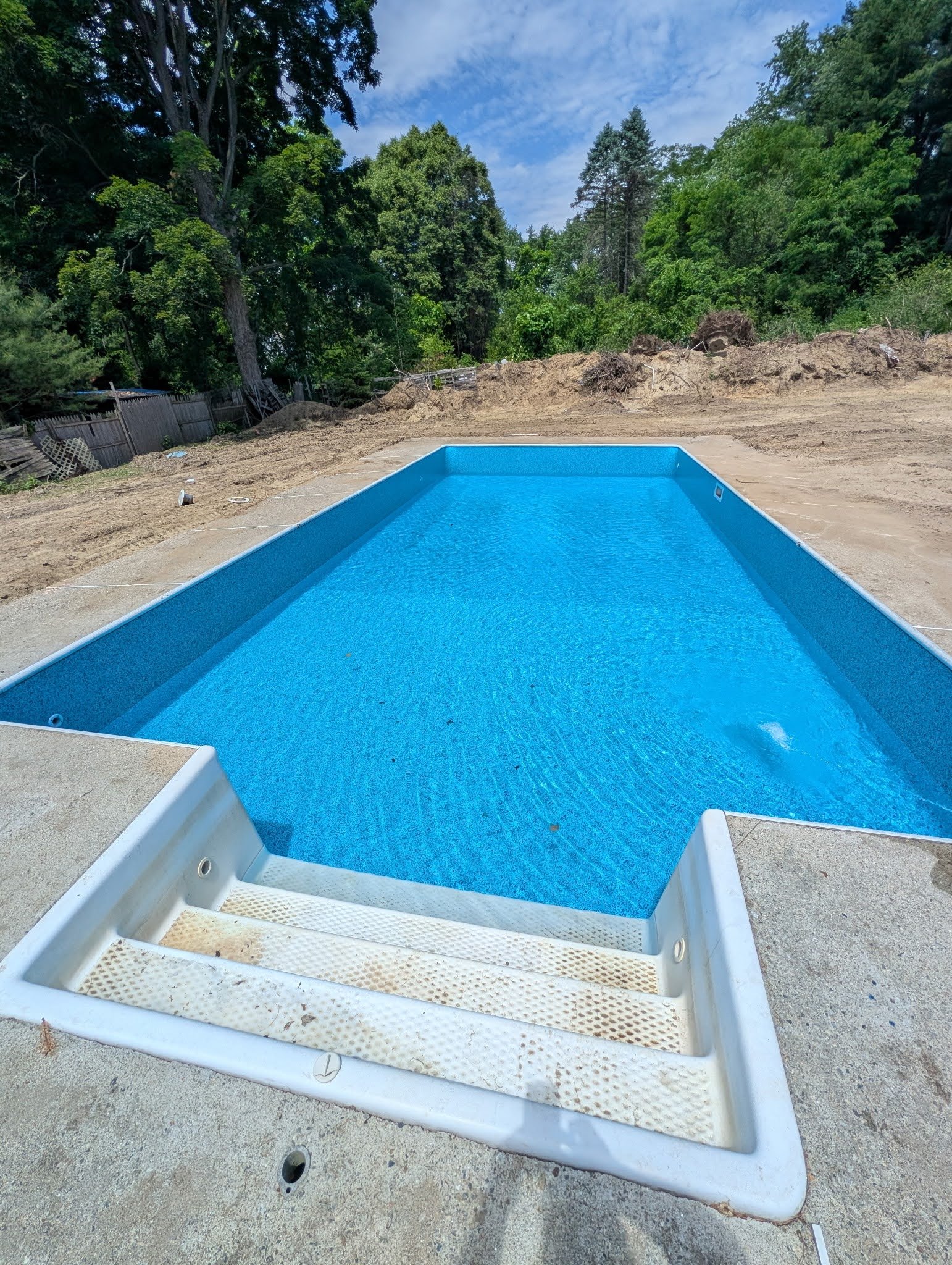 Empty swimming pool on concrete deck in backyard with dirt and trees in background.