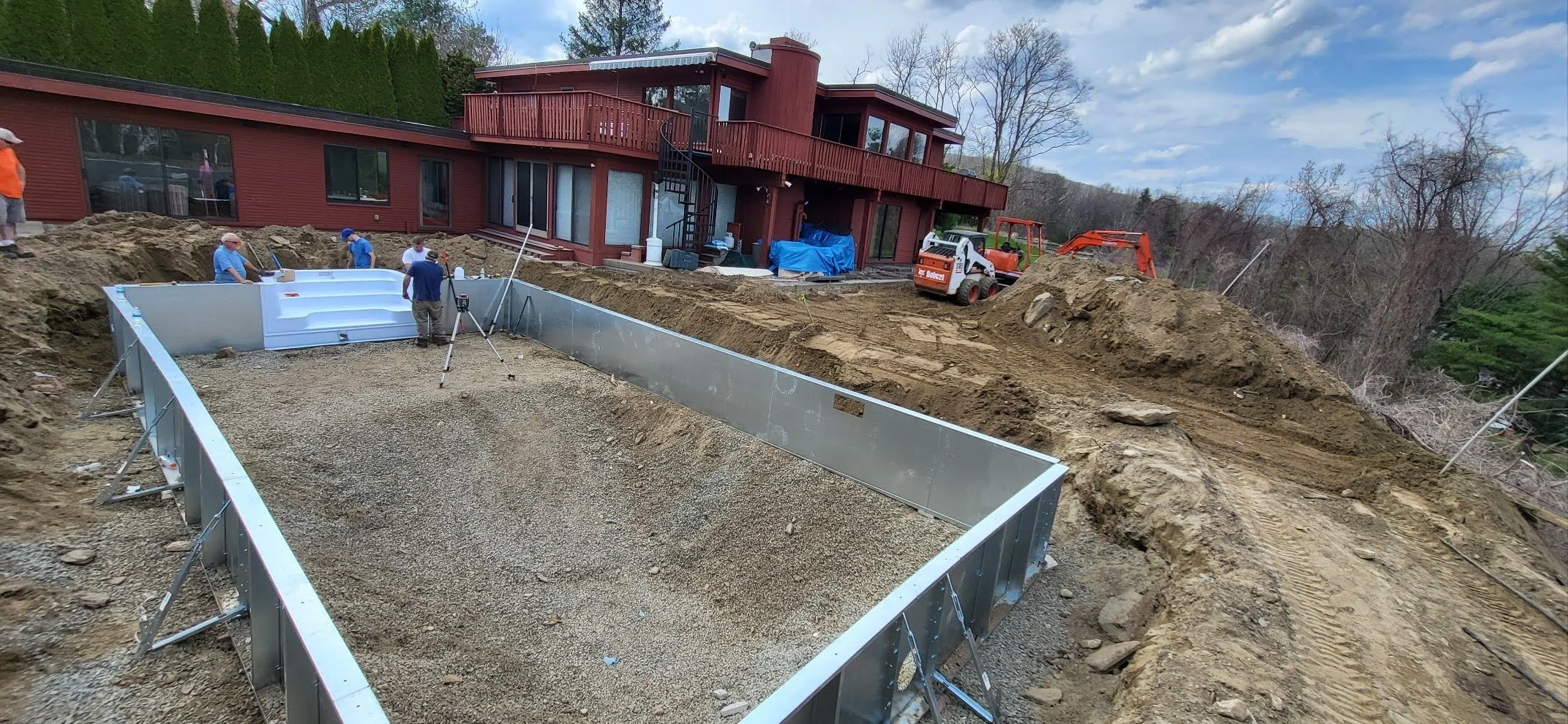 Construction site with workers installing a pool foundation in front of a red house, machinery, and dirt mounds under a cloudy sky.