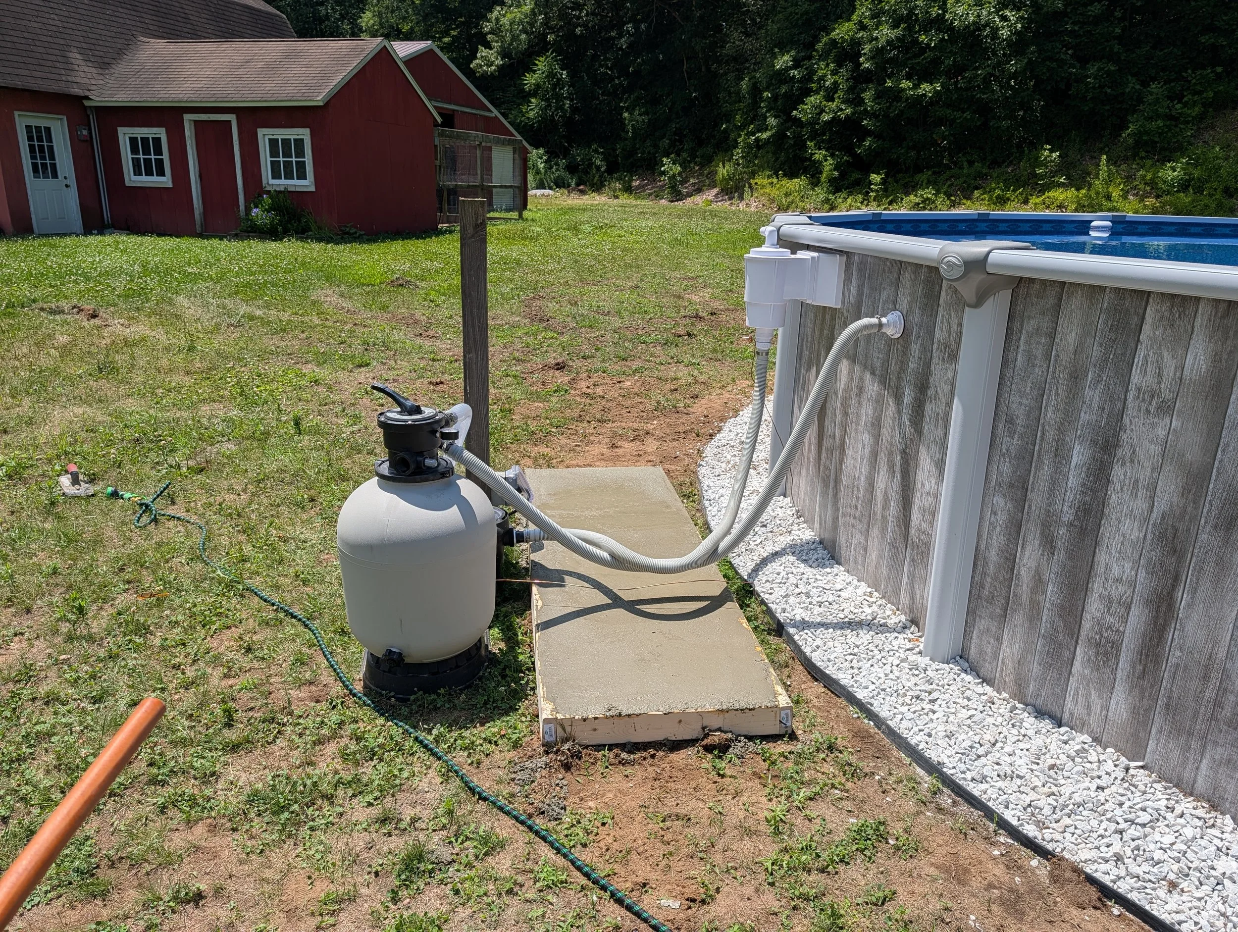 An above ground swimming pool with a white exterior and blue interior water, connected to a filter system, on a concrete pad in a backyard. There is a red barn with white trim and several windows in the background, surrounded by green grass and trees