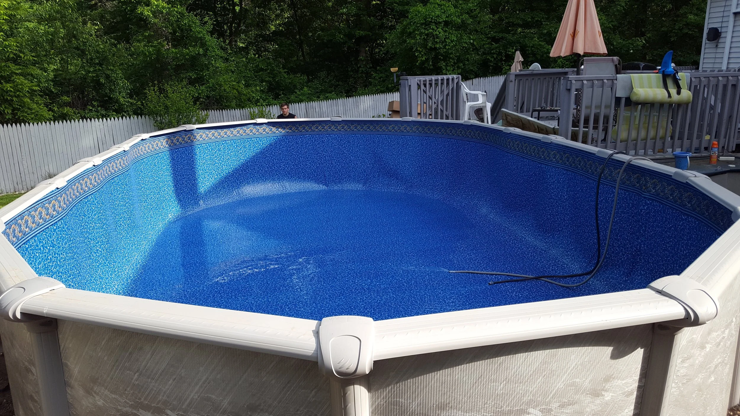 In-ground swimming pool with a blue interior, located in a backyard with a white fence and green trees. Part of a deck with outdoor furniture and an umbrella is visible in the background.