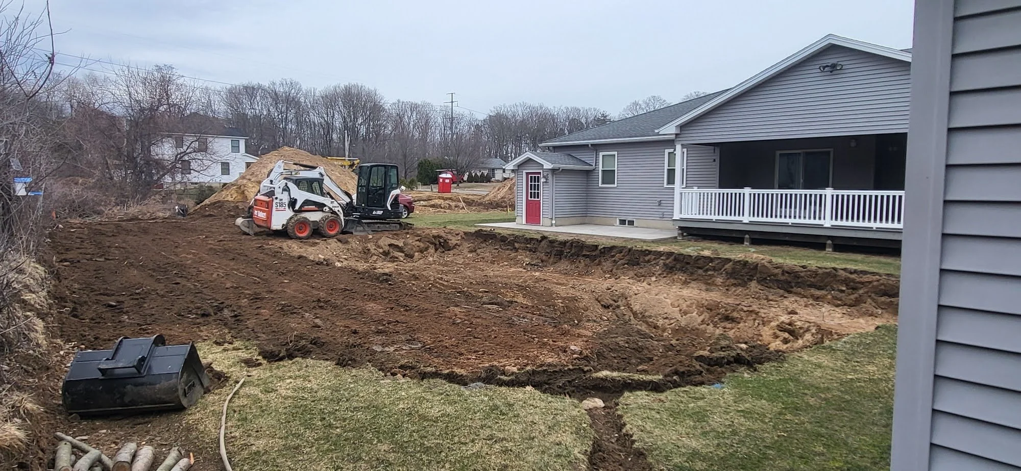 A residential backyard with a construction site where the ground has been excavated and a small skid-steer loader parked nearby. There is a house with gray siding and a porch on the right side, and a pile of dirt and construction equipment in the bac