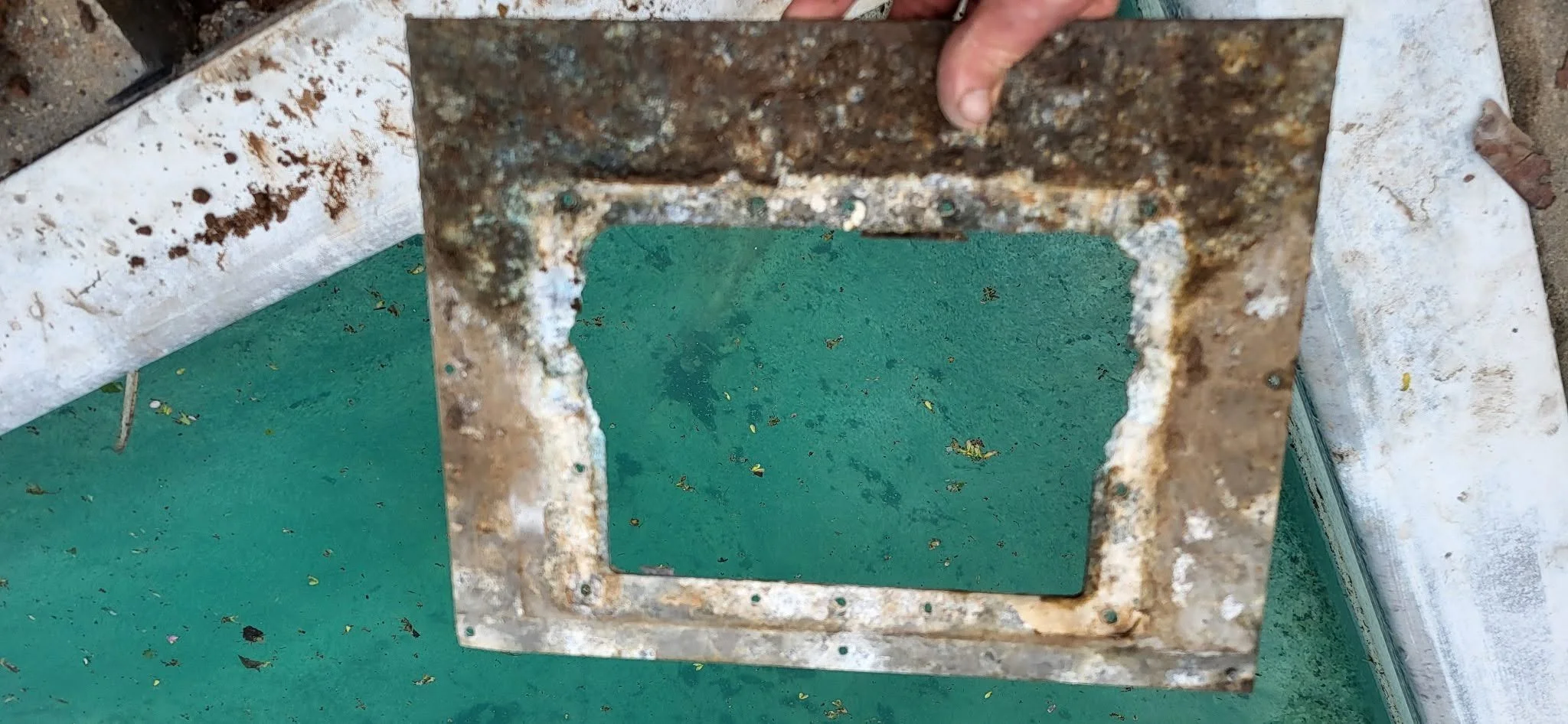 A close-up of a rusty metal object with a rectangular hole, held by a person's hand, placed on a green surface with debris and other objects around.