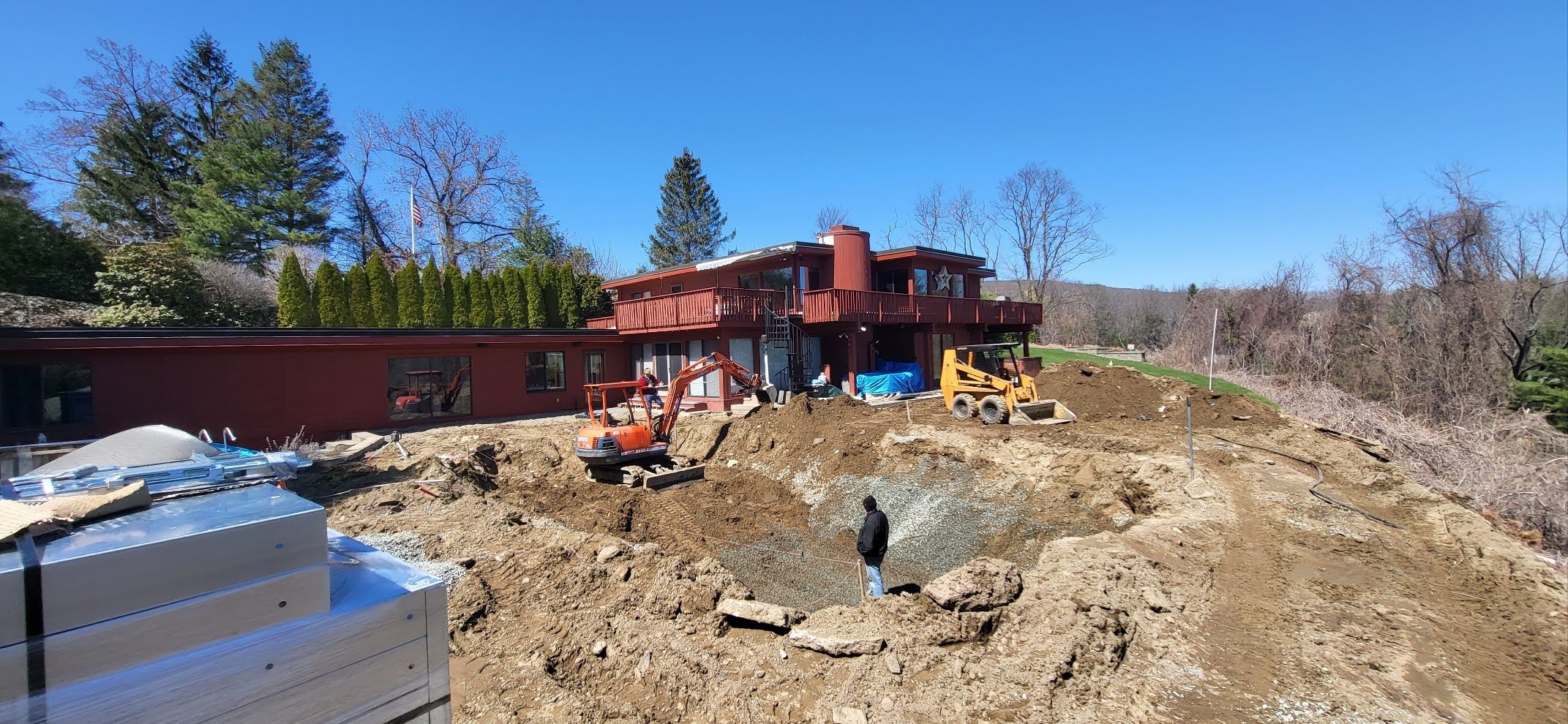 Construction site with a house in progress, construction equipment, and workers, on a sunny day with clear blue sky and trees in the background.