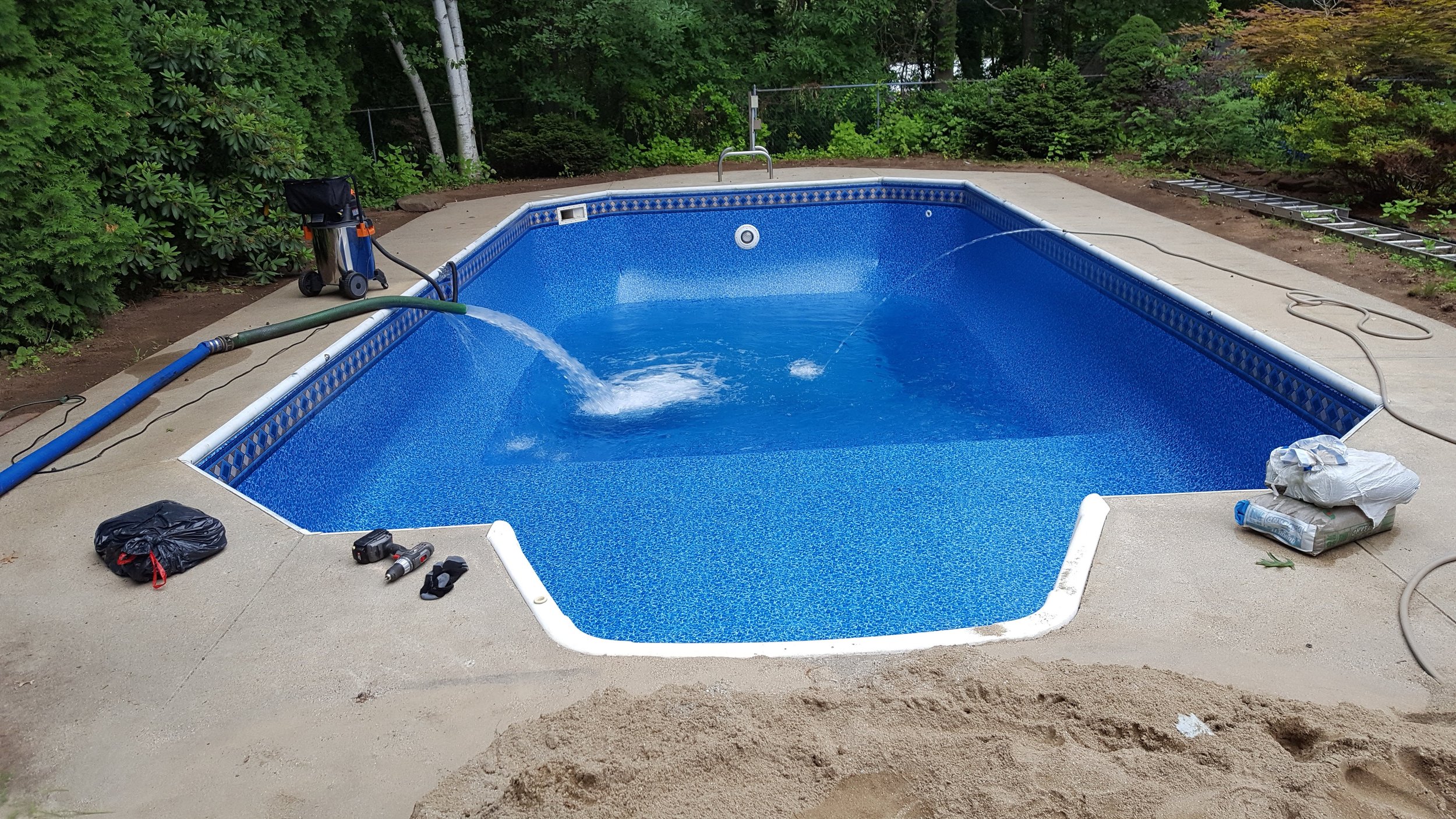 In-ground blue swimming pool with partially filled water, surrounded by a concrete deck, construction tools and equipment nearby, and lush green trees in the background.