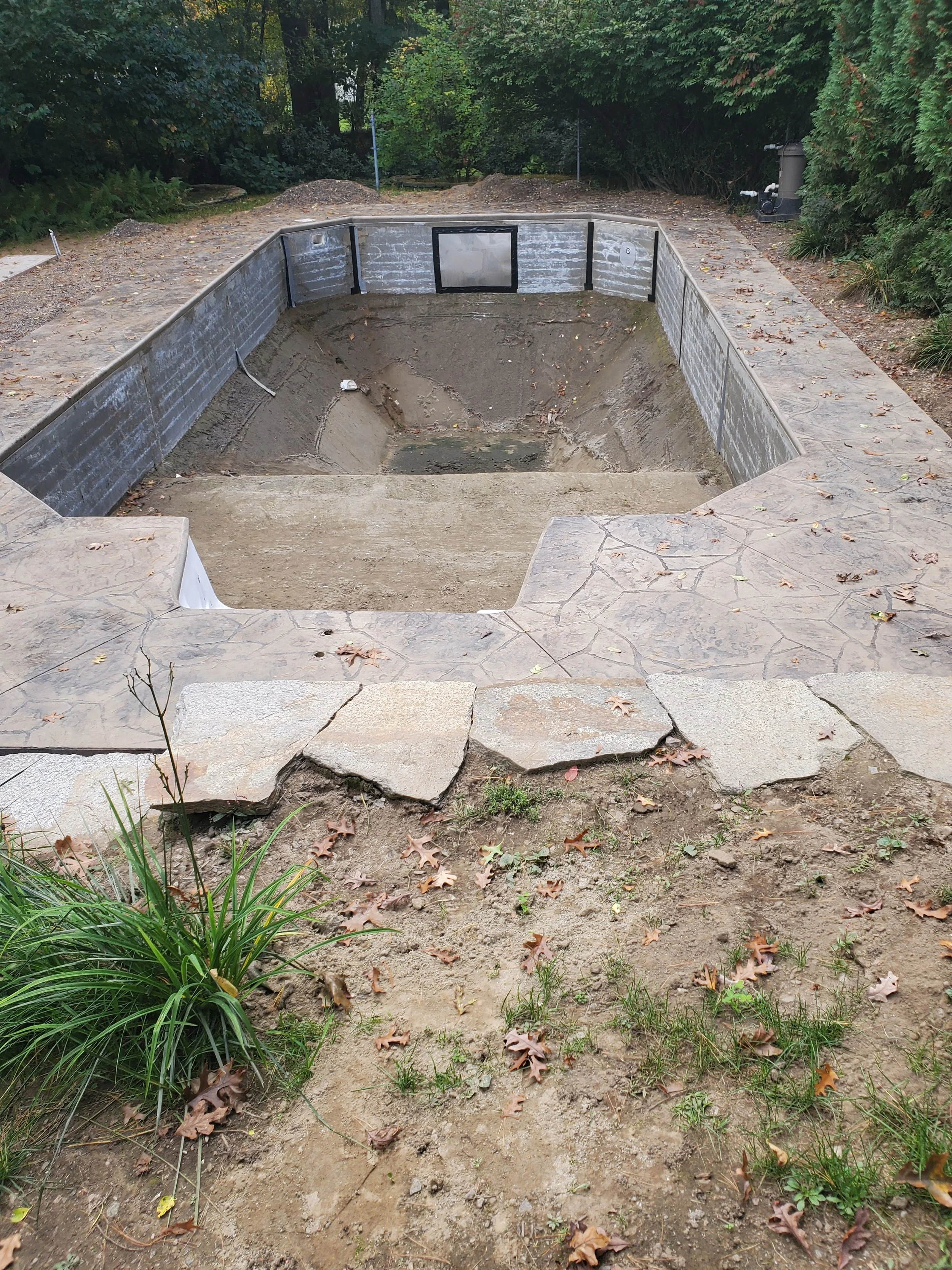 A backyard pool under construction with the liner partially installed, surrounded by stone paving and grass, with trees and bushes in the background.