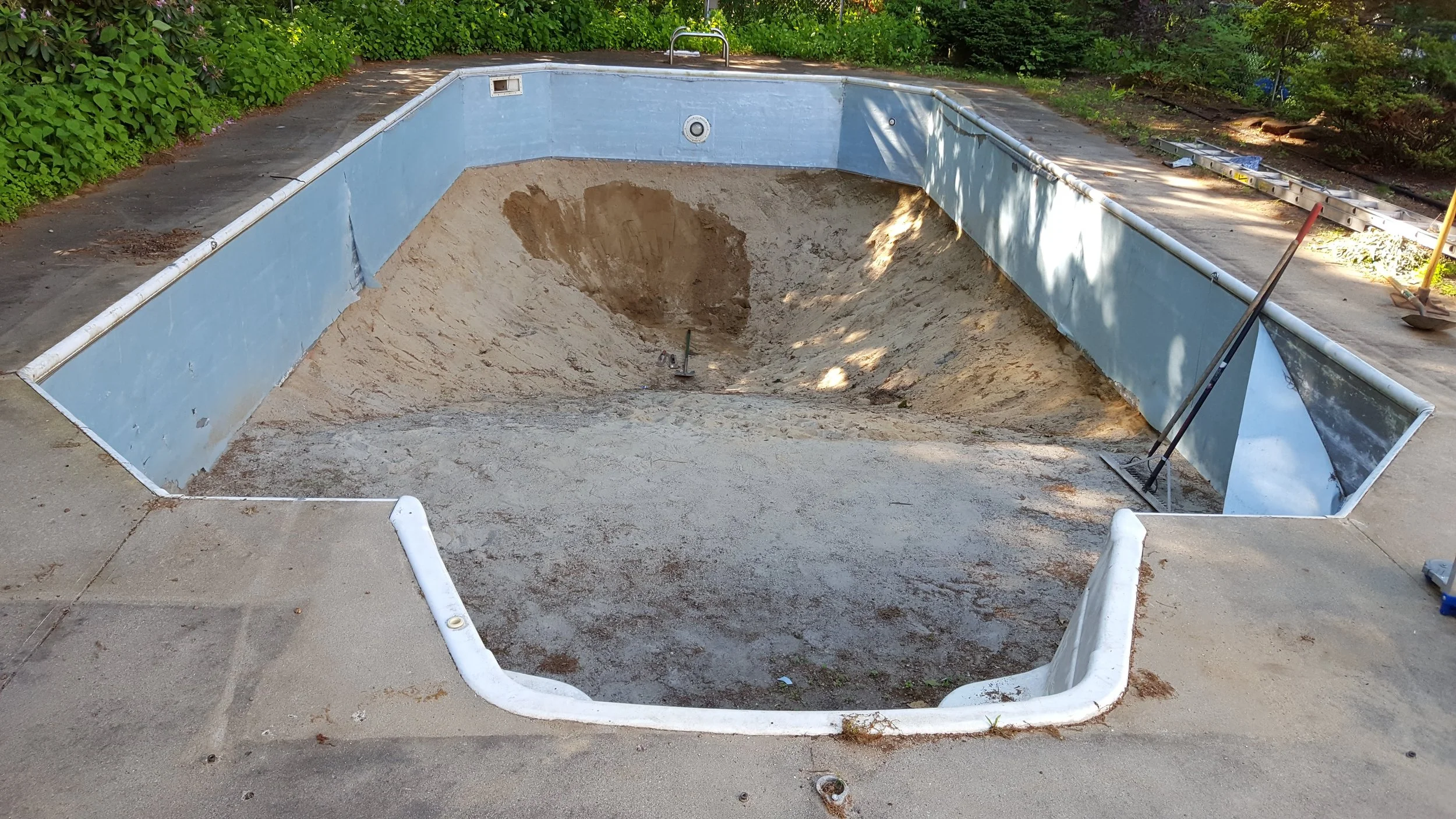 Empty swimming pool under undergoing renovation with sand and dirt inside, surrounded by tools including shovels and a ladder, with overgrown bushes and trees in the background.
