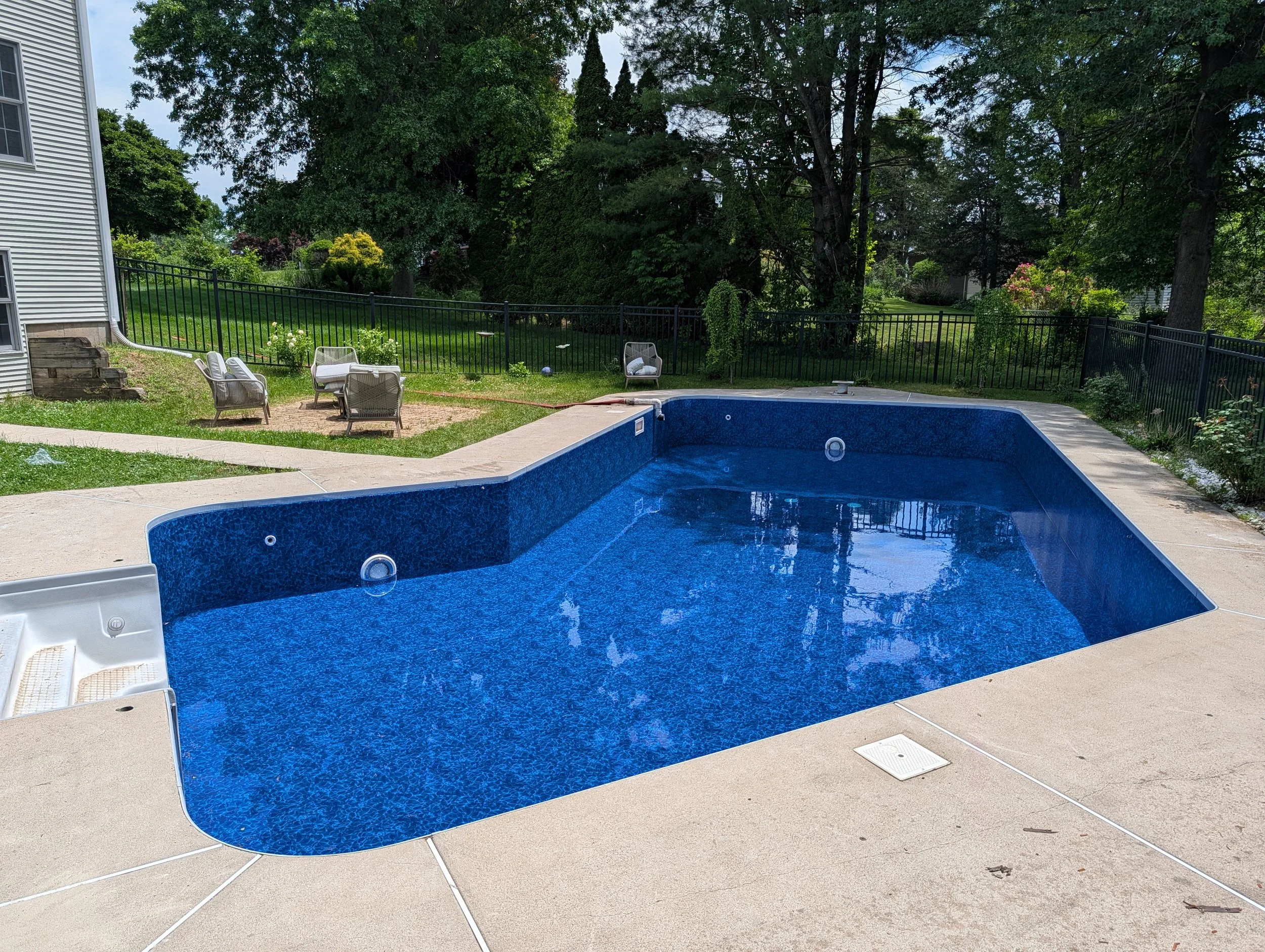 A backyard with an in-ground blue swimming pool, surrounded by a concrete patio with white lounge chairs. There is a black metal fence around the yard, with trees and greenery in the background and a house wall on the left side.