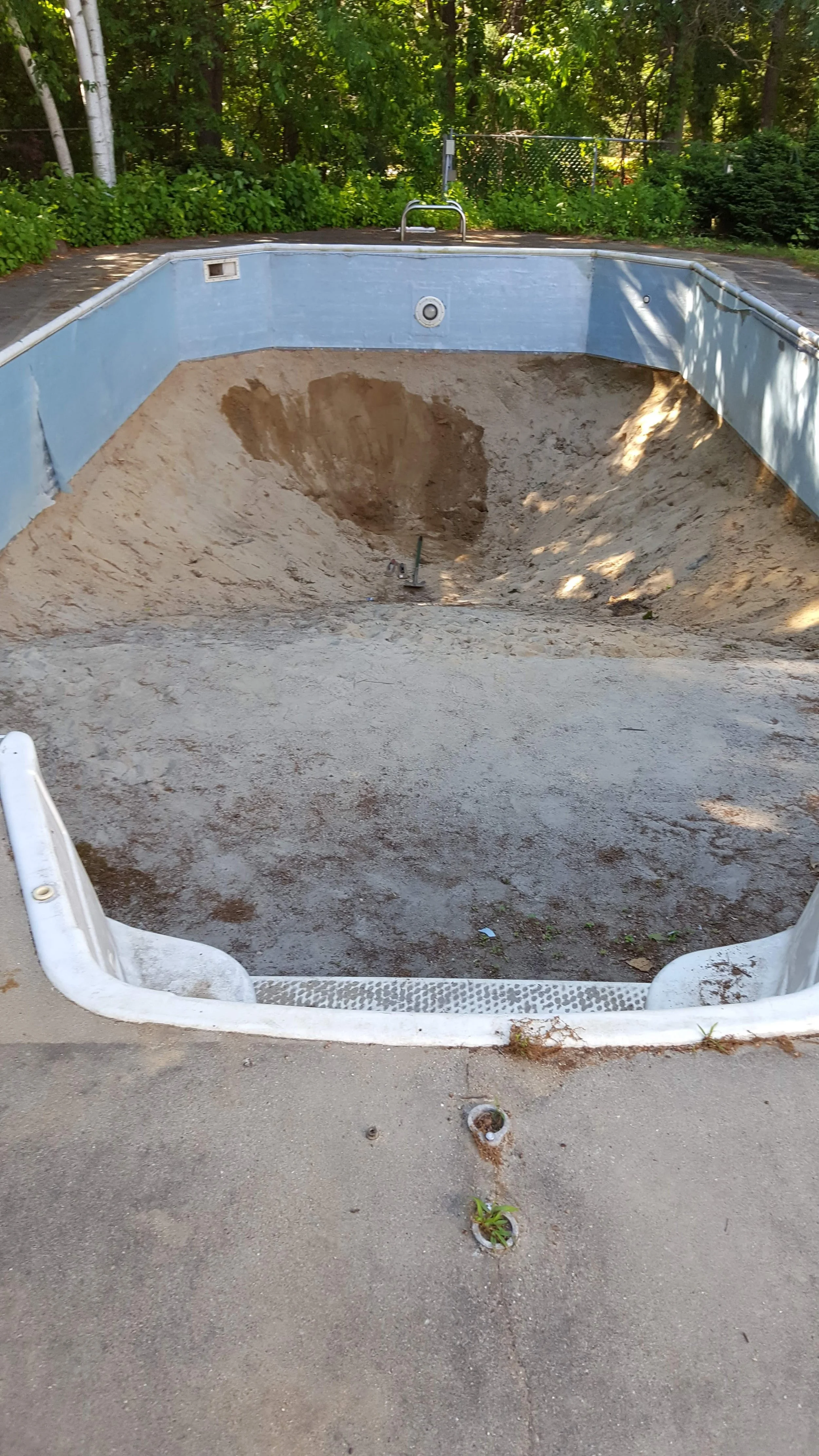 Empty swimming pool with sandy bottom, surrounded by a concrete deck and green trees and bushes in the background.