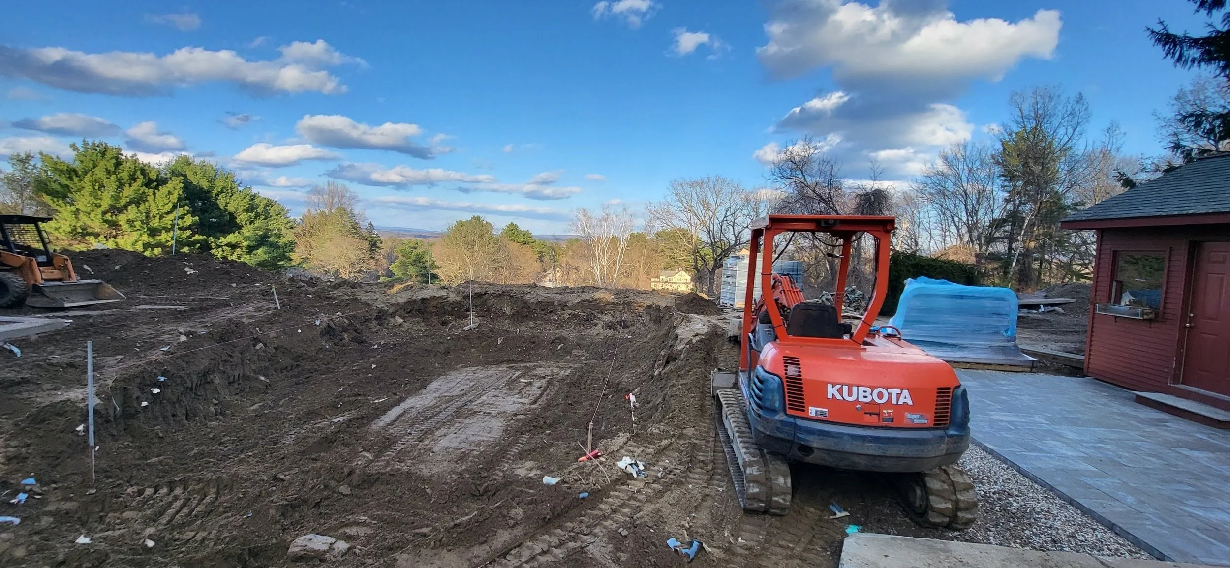 A construction site outdoors with a small orange Kubota mini excavator, dirt piles, and construction materials. The background includes trees, a blue sky with clouds, and a distant view of houses.
