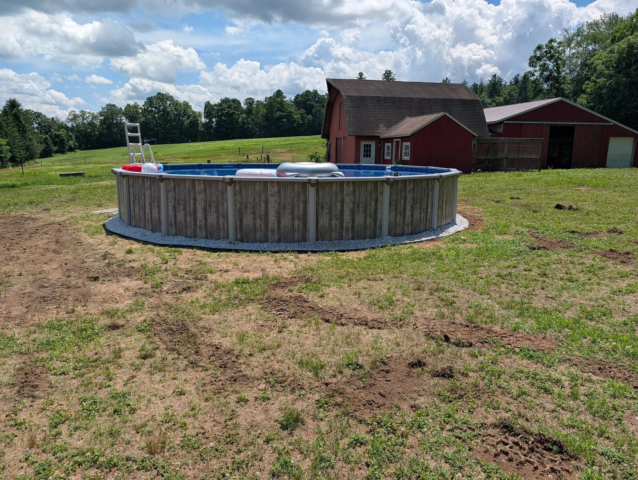 An above ground swimming pool with a ladder, a float, and a small toy in a grassy backyard next to a red barn, under partly cloudy skies.