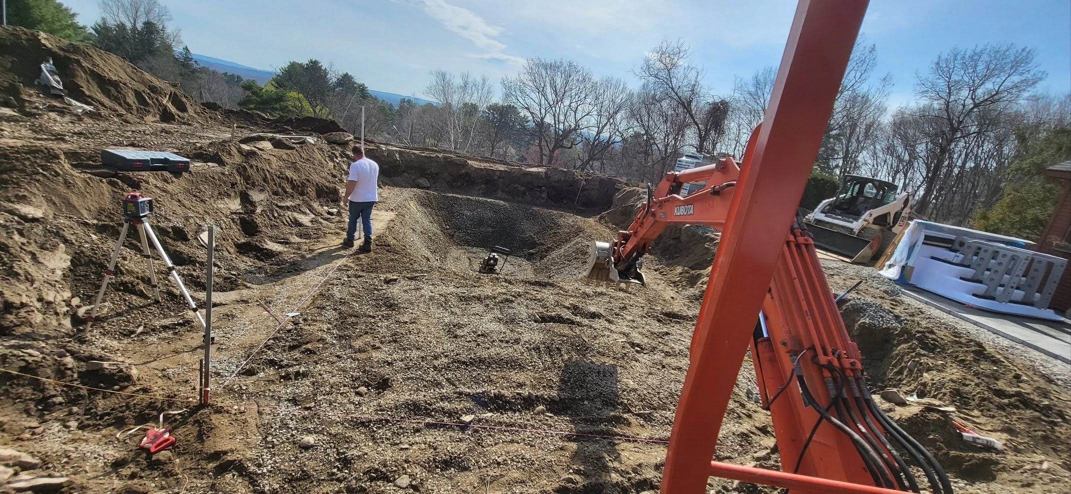 Construction site with excavator and survey equipment, a worker standing on dirt, trees and blue sky in the background.