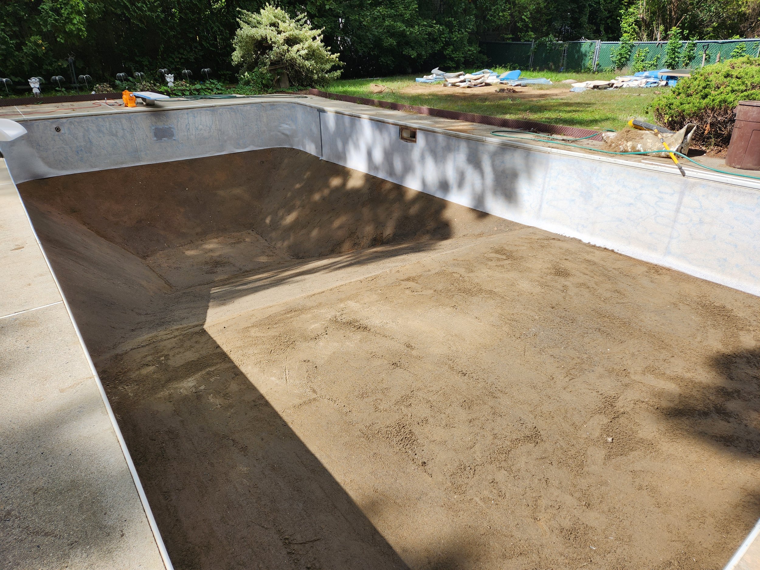 Empty swimming pool under construction with dirt and sand inside, surrounded by greenery, tools, and construction materials.