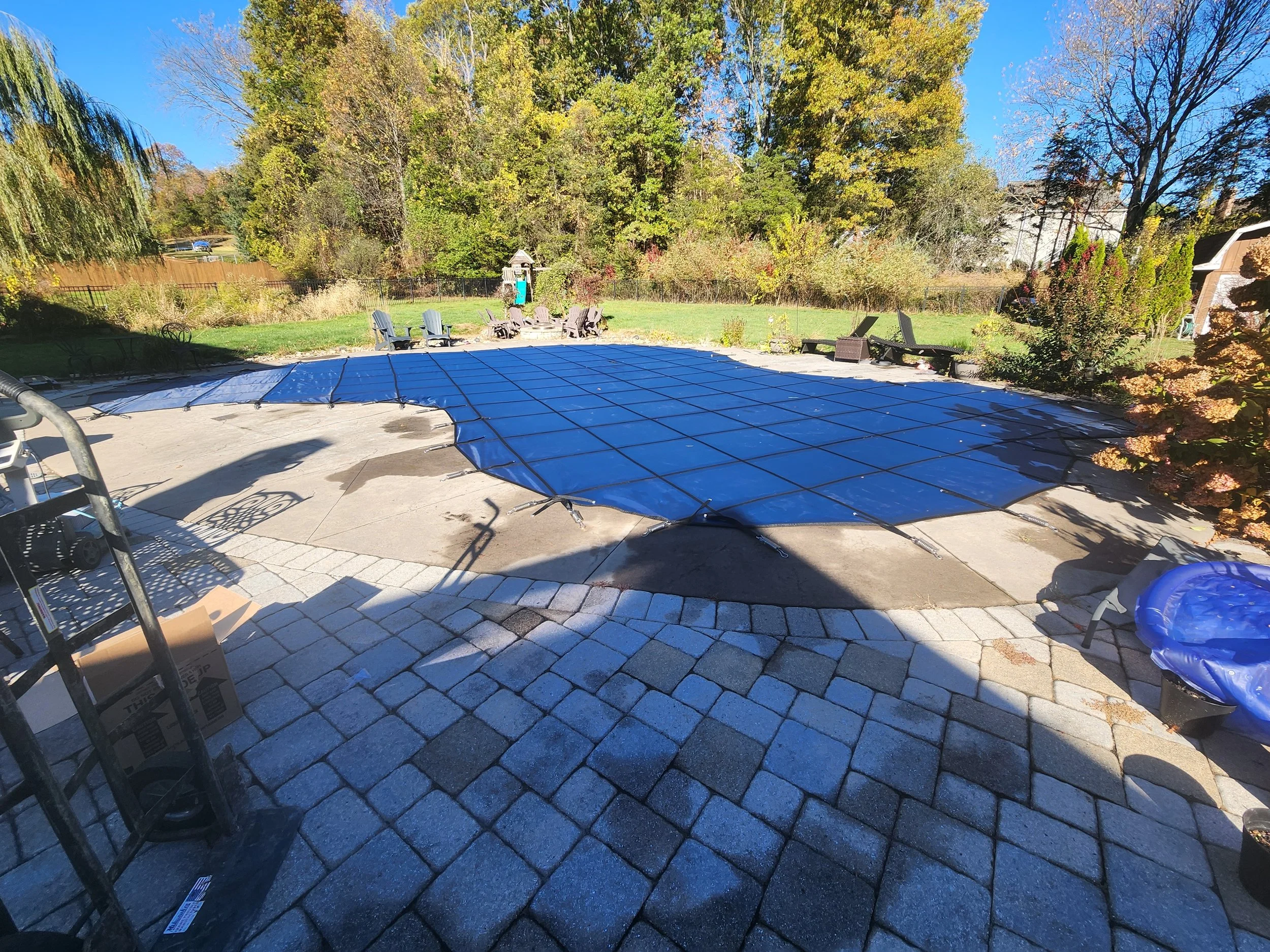 Backyard with newly installed paver patio, surrounded by trees with fall foliage, some chairs and a covered pool or hot tub.