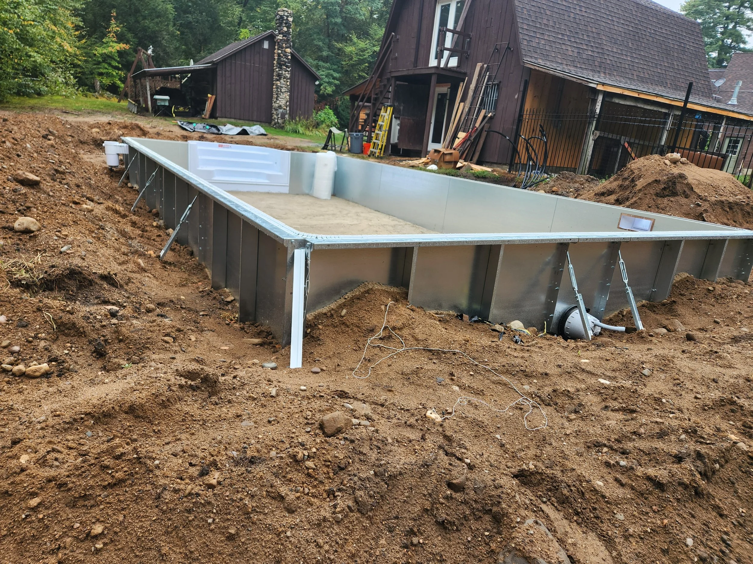 An outdoor construction site where a pool is being installed, with dirt and soil surrounding it and a house with brown wooden siding in the background.