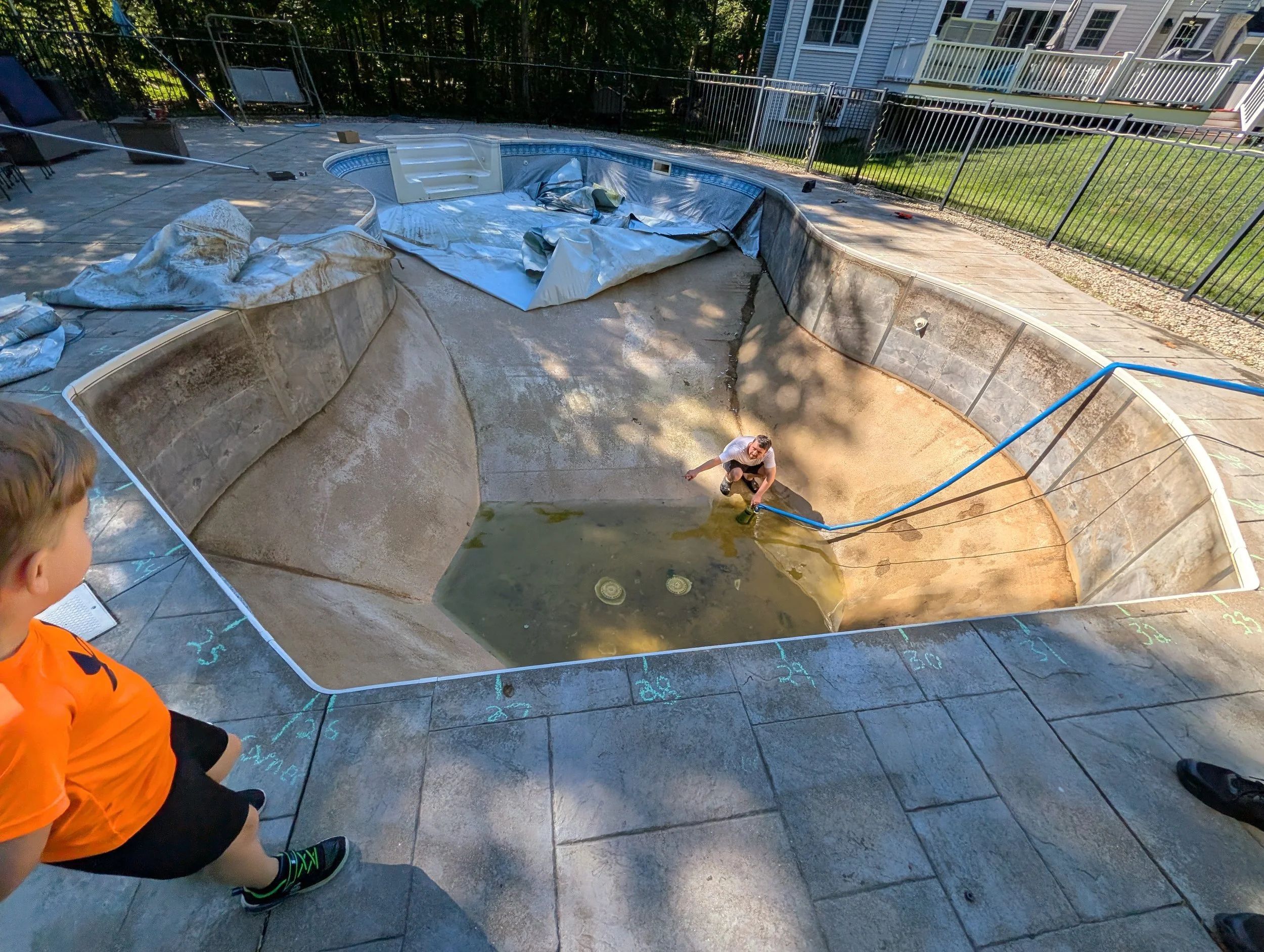 People working on the construction or renovation of a swimming pool in a backyard. The pool is empty, with a blue tarp or liner partially installed. Chalk marks and numbers are written on the surrounding concrete surface.