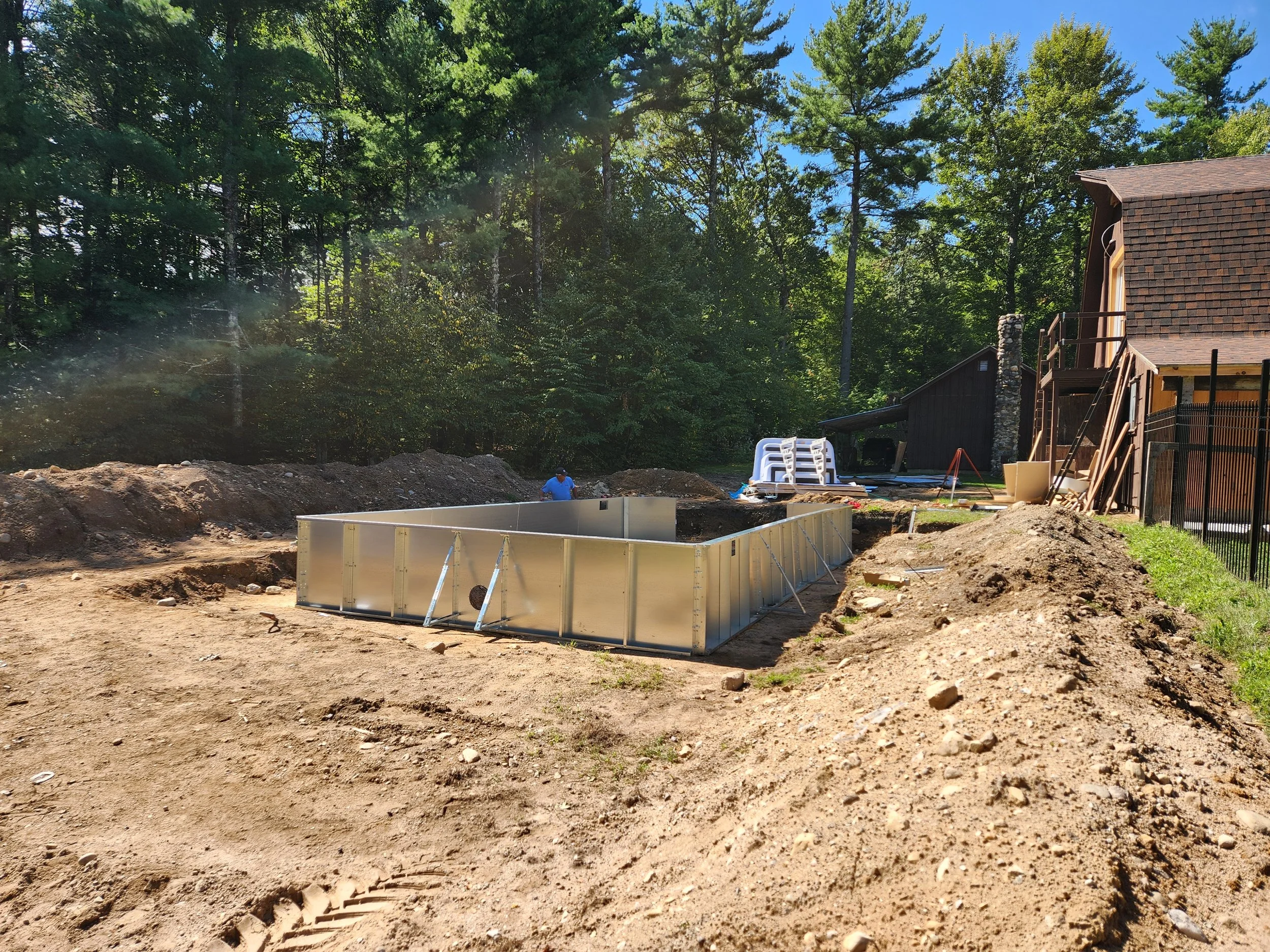 Construction site with a partially built pool, dirt ground, and construction materials. A person in blue is working near the pool, with trees and a house in the background.