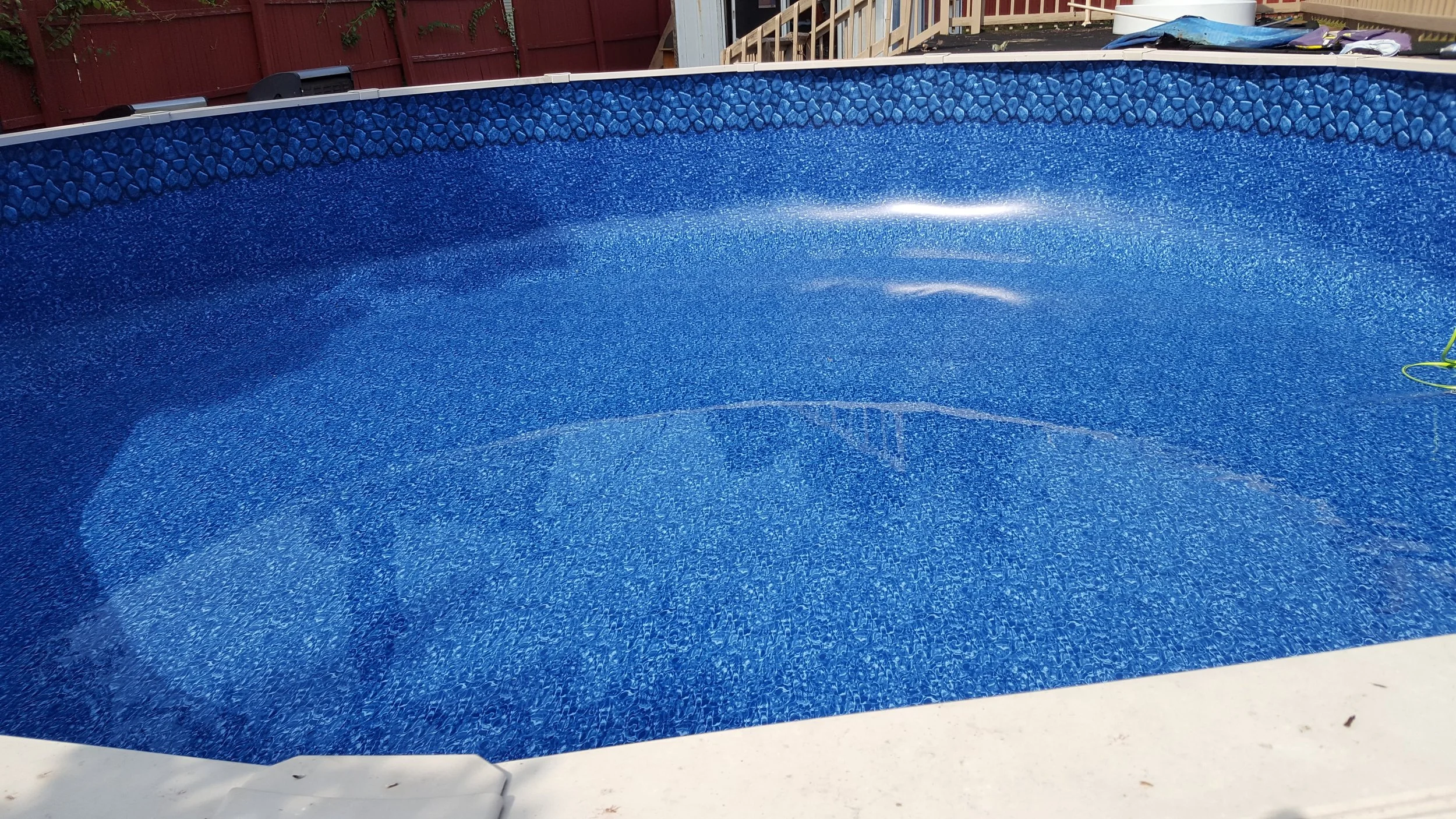 Empty swimming pool with blue tiling, surrounded by a concrete deck and a red wooden fence in the background.