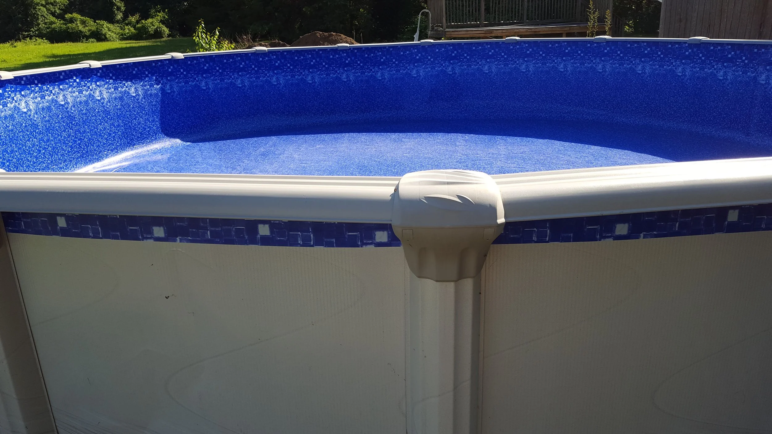 Above ground swimming pool with blue mosaic tile interior, set outdoors on grass with trees and a wooden deck in the background.