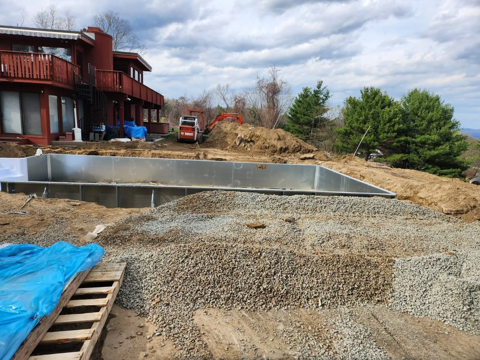 Construction site with an in-ground pool frame, gravel pile, construction equipment, and a red house with a balcony in the background.