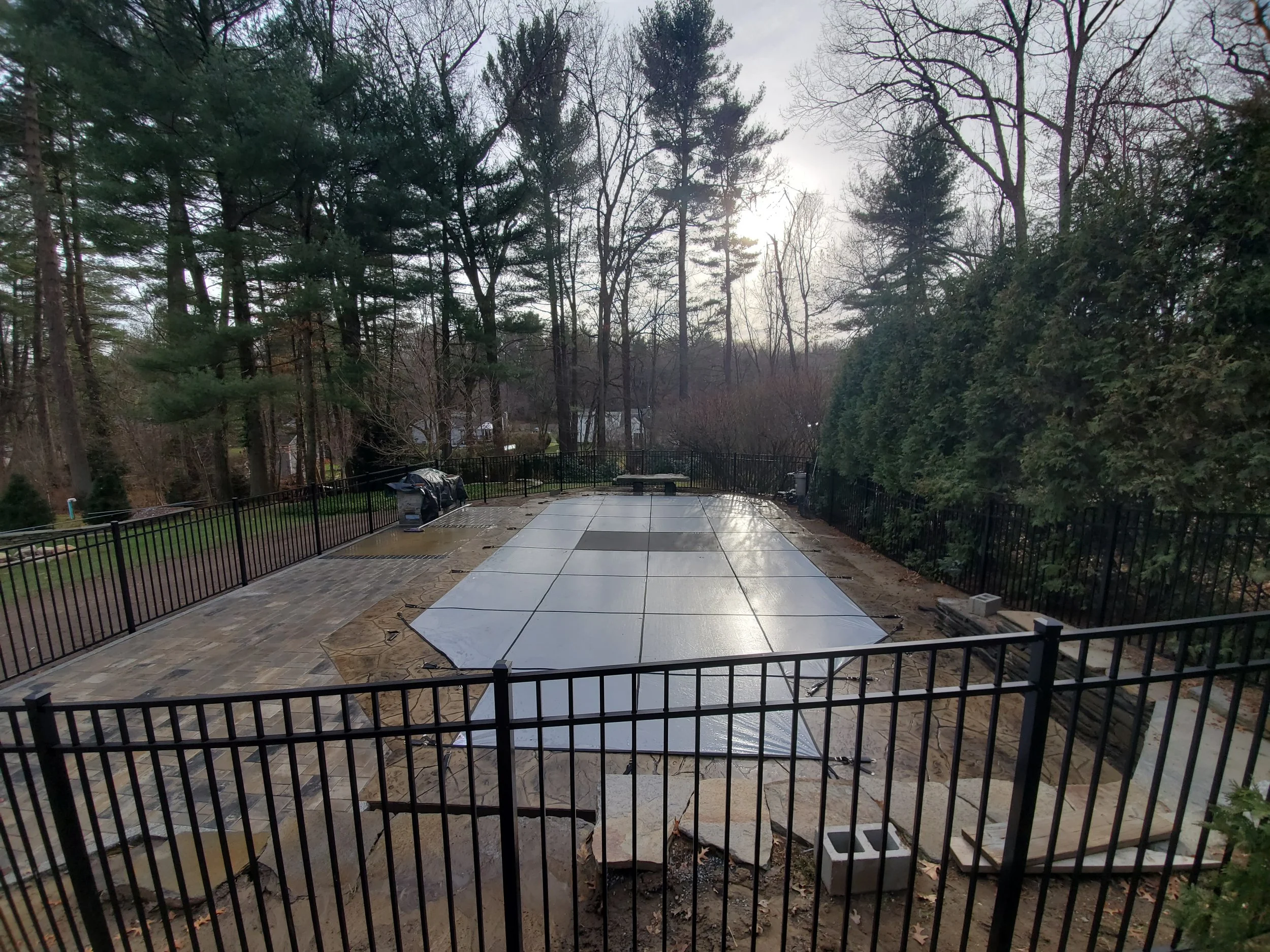 A backyard with a patio area under construction, surrounded by a black metal fence, with trees in the background and an overcast sky.