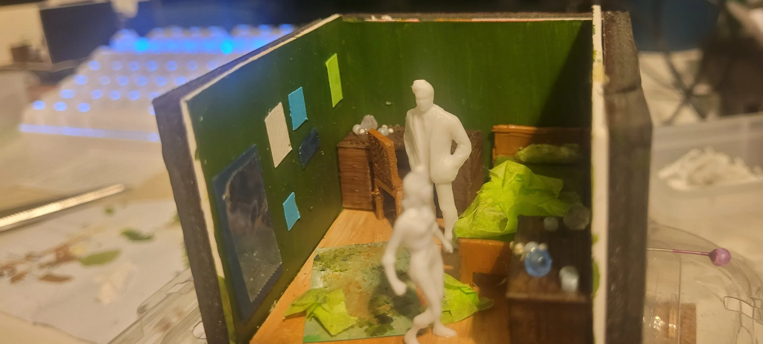 Lexy Kimler's miniature diorama of a room with a man figure standing, surrounded by furniture and decorations, painted green walls with pictures, and paper leaves on the floor.