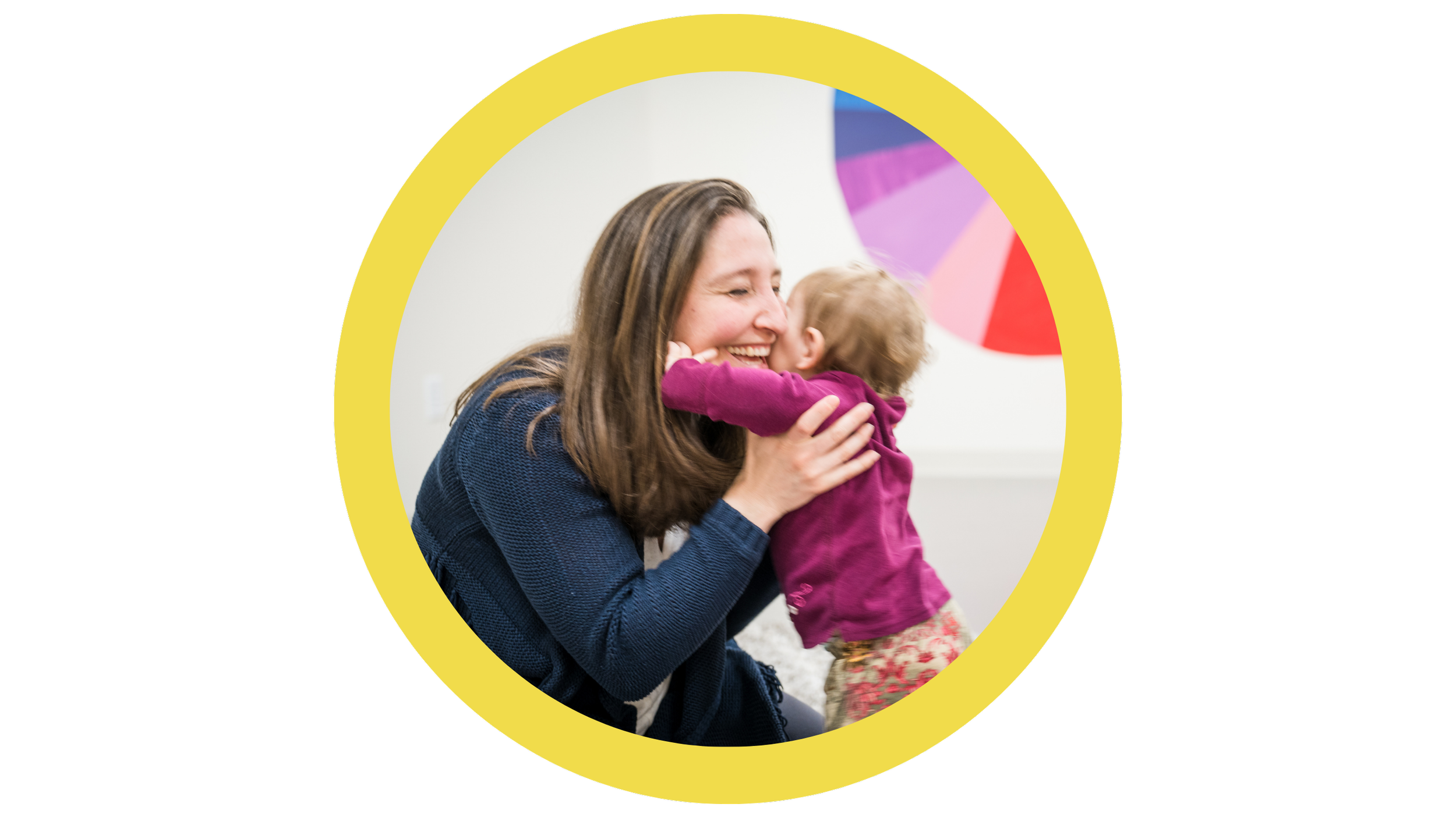 A woman with long brown hair smiling and hugging a young girl with blonde hair, wearing a purple top, in a bright room with colorful wall art in the background.