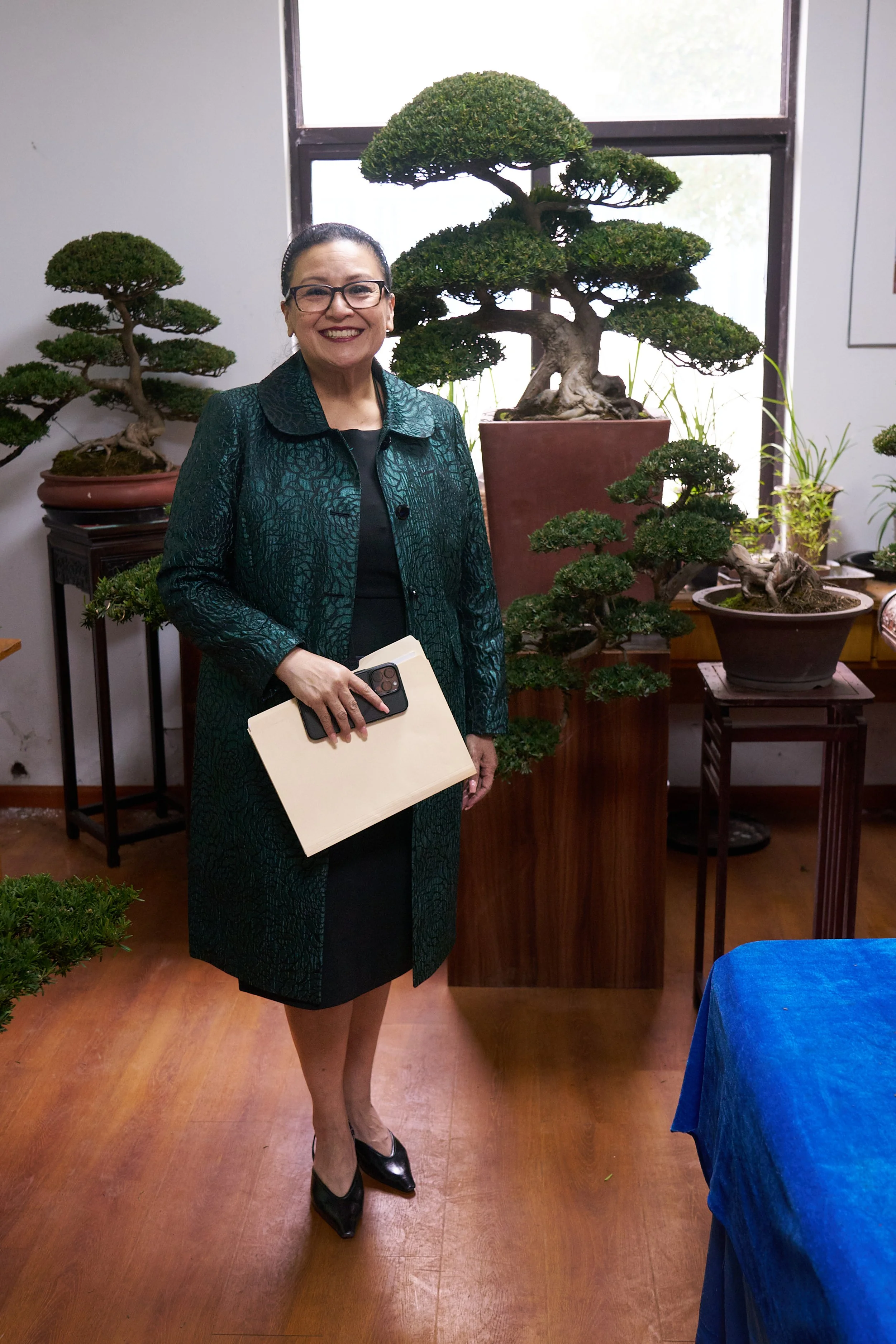 A woman in a dark teal patterned jacket, black dress, and black heels standing inside near a large bonsai tree and smaller bonsais, holding a notebook and a smartphone, with natural light coming through a window.