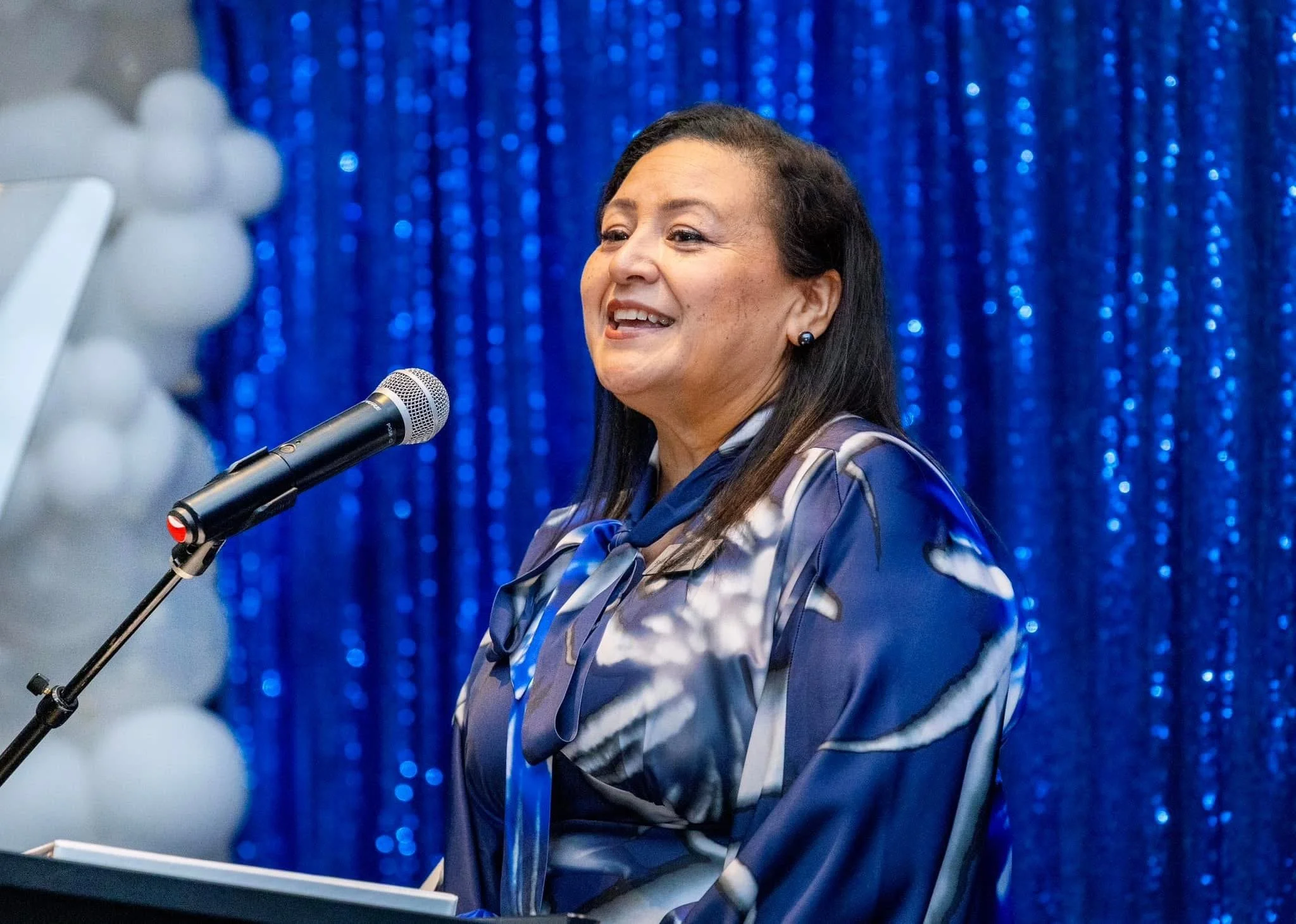 A woman speaking into a microphone on stage with a blue, shimmering curtain background.