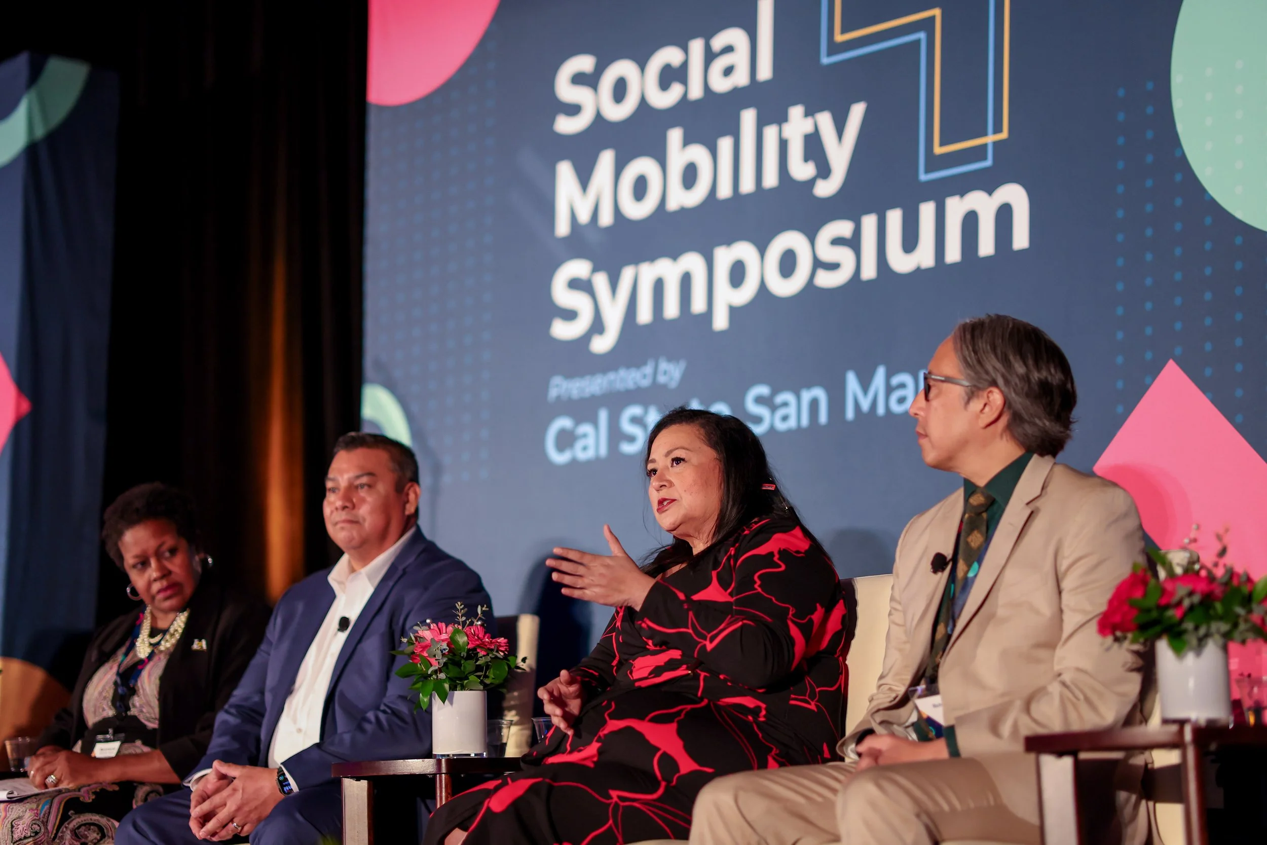 A panel of five diverse individuals seated at a discussion at the Social Mobility Symposium, with a large screen behind them displaying the event's name.