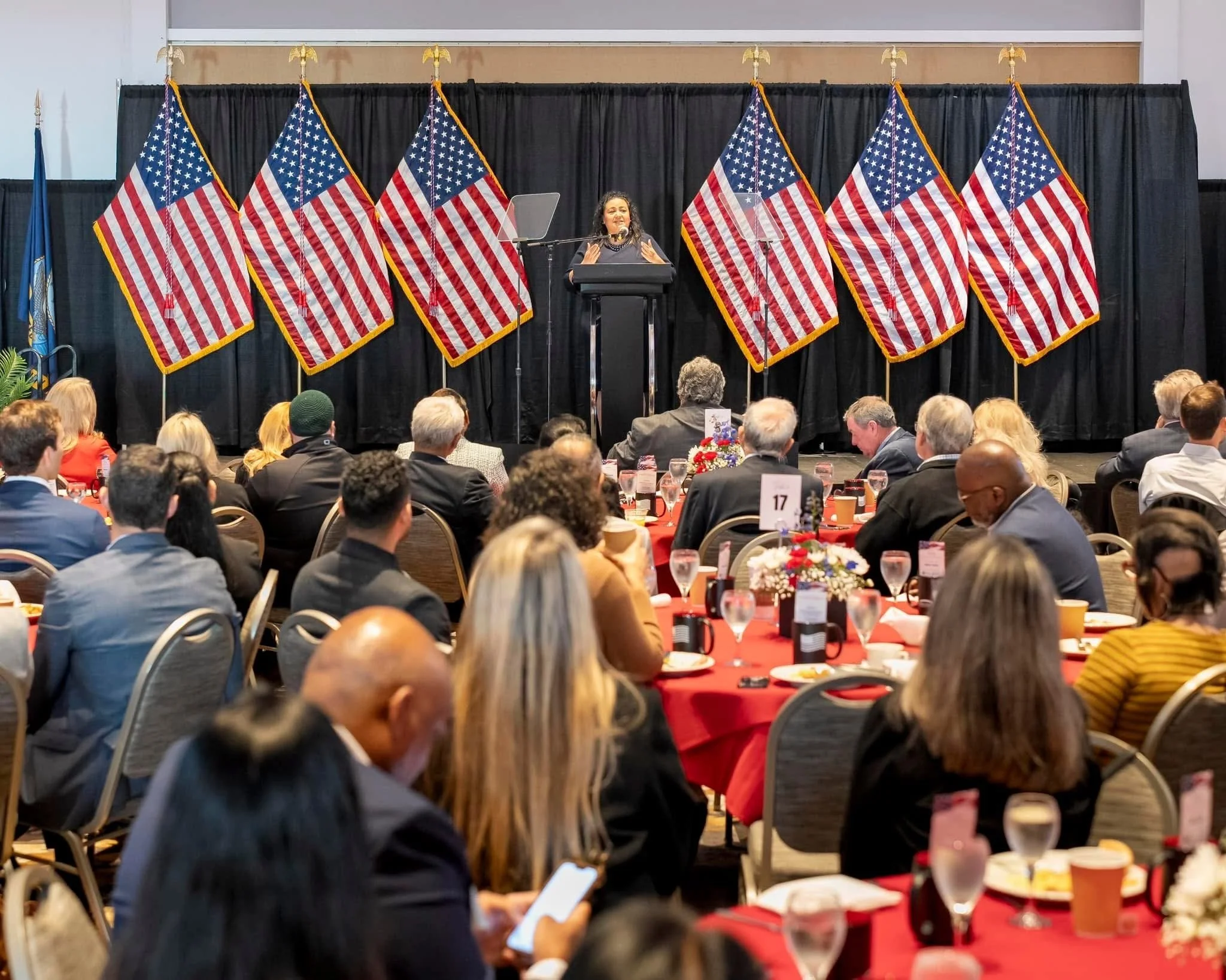 A woman speaking at a podium during a formal event with multiple American flags in the background. Audience members are seated at tables with red tablecloths and floral centerpieces.