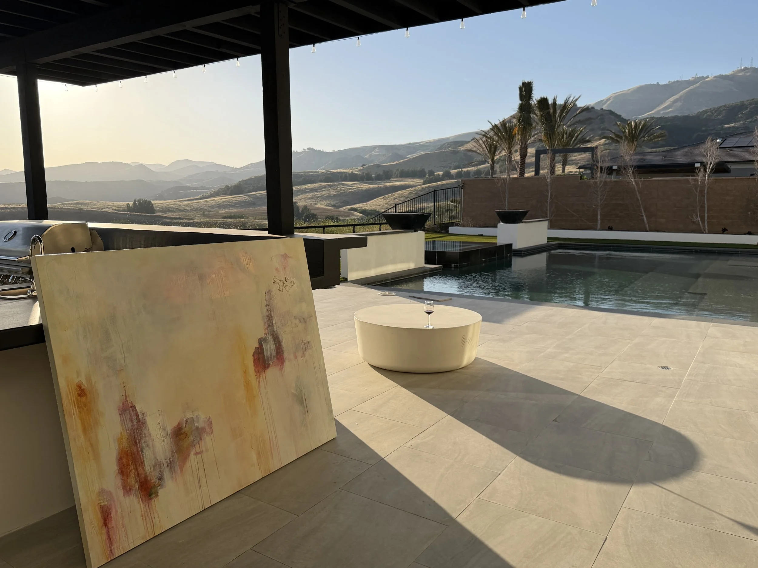 A patio area with a swimming pool, modern chairs, and a table with a glass of wine, overlooking mountains and palm trees in the background.