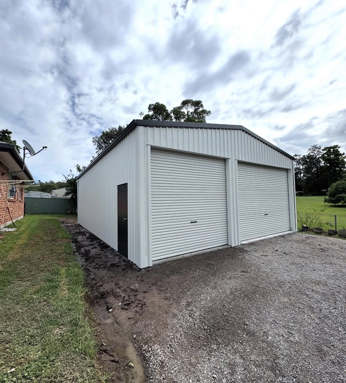 A small white metal building with two roll-up garage doors and a black side door, situated on a gravel driveway with patches of mud, in a suburban backyard with green grass and trees under a partly cloudy sky.