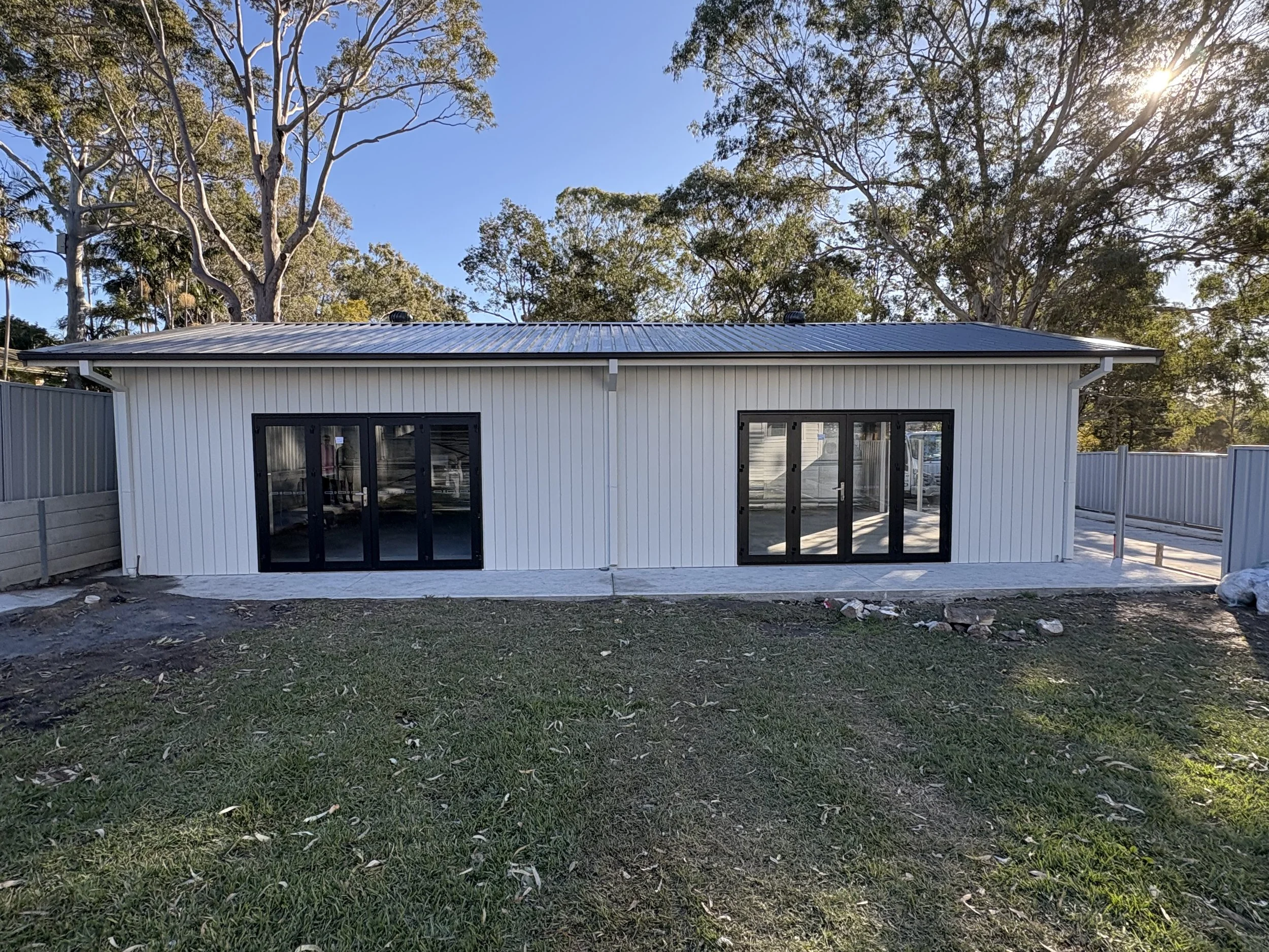 A modern house with large sliding glass doors, white exterior siding, and a metal roof, situated in a backyard with a grass lawn and tall trees in the background under a clear blue sky with the sun shining.